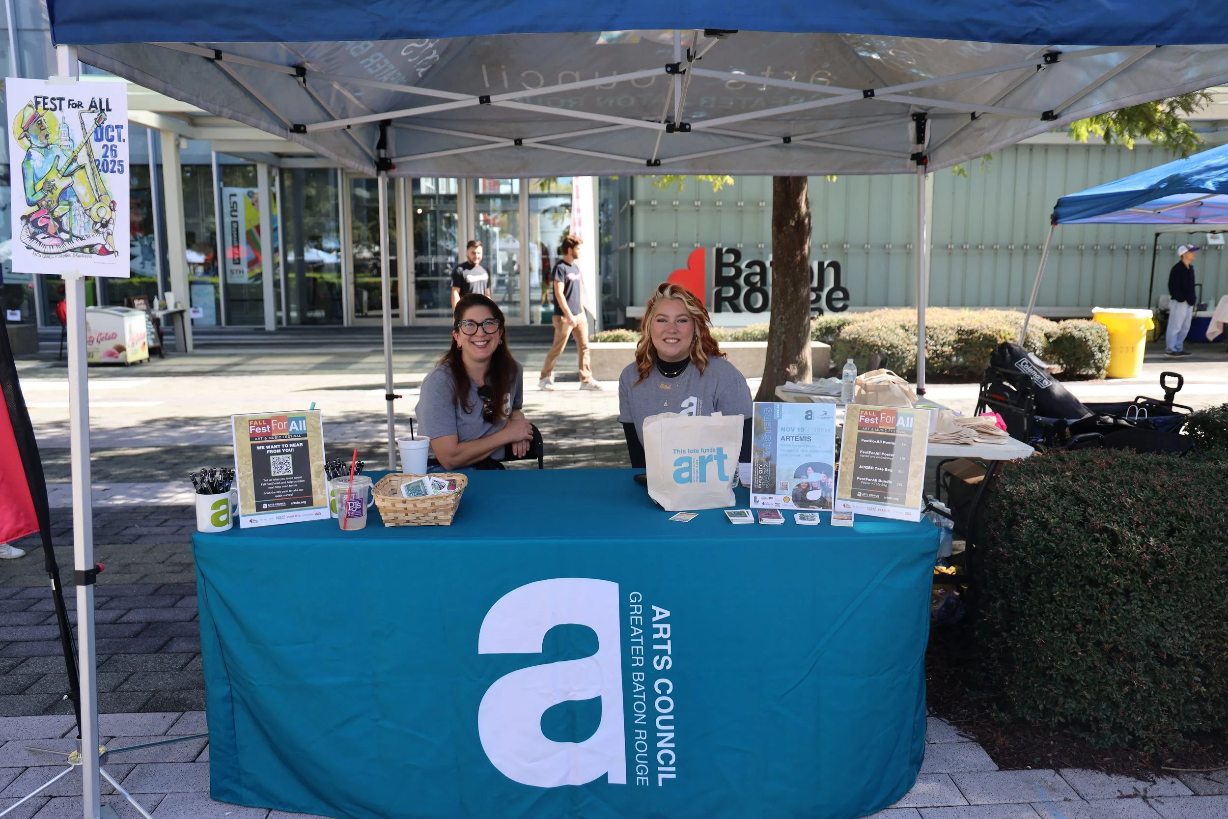 Two women sitting behind a table at an outdoor event booth, with signs promoting an art fund and festival, under a canopy tent on a city sidewalk.