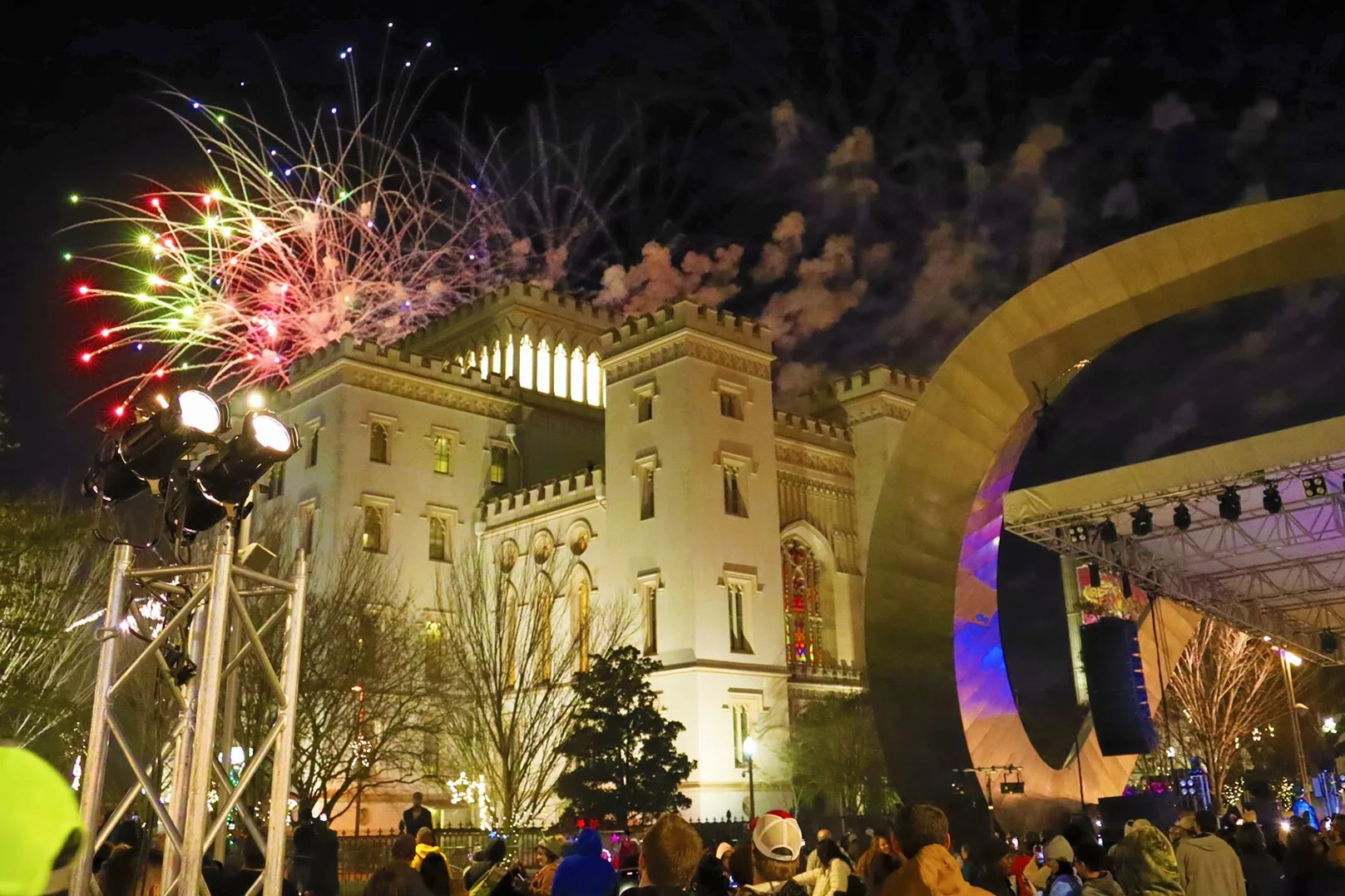 Fireworks light up the night sky above a castle-like building at an outdoor event with a stage and many spectators.