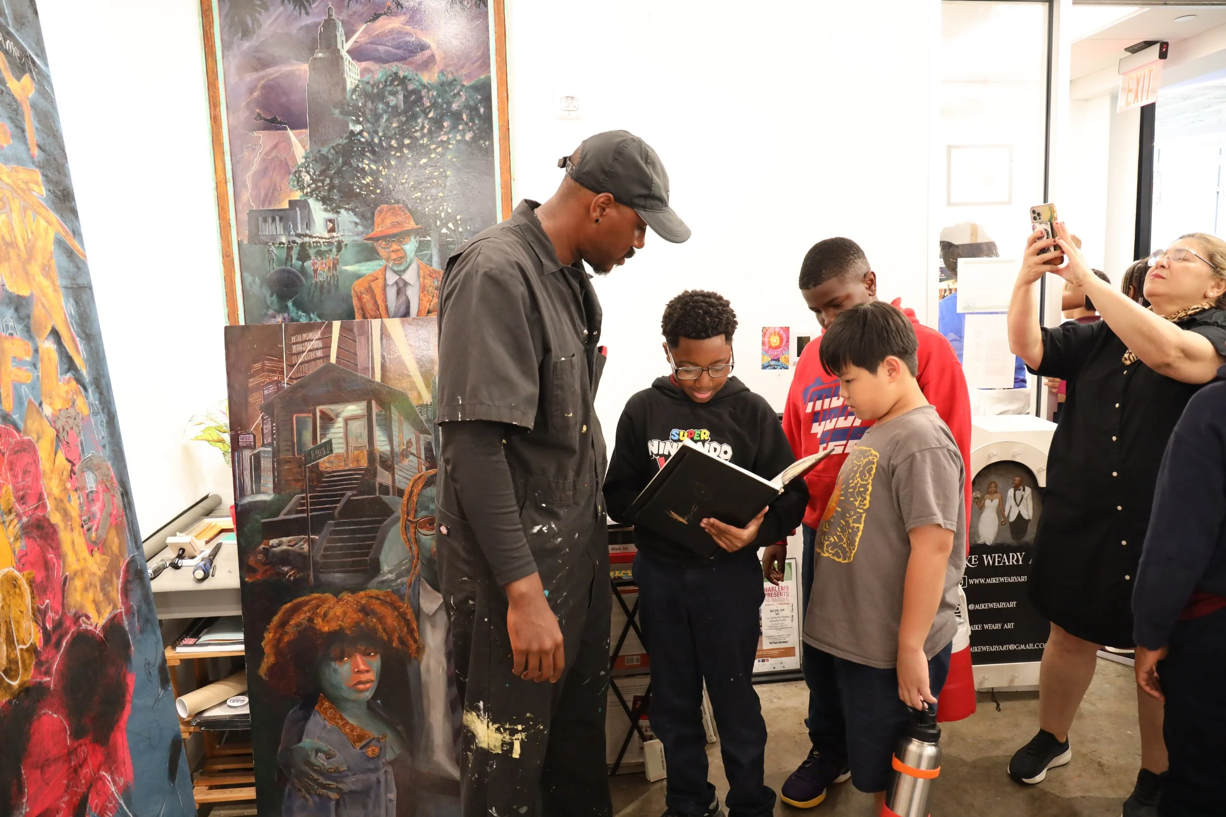 A group of young boys and an adult man are gathered in an art gallery, looking at artwork displayed on the walls and tables. A woman on the right is taking a photo with her phone. The gallery features vibrant paintings, including portraits and abstract art.