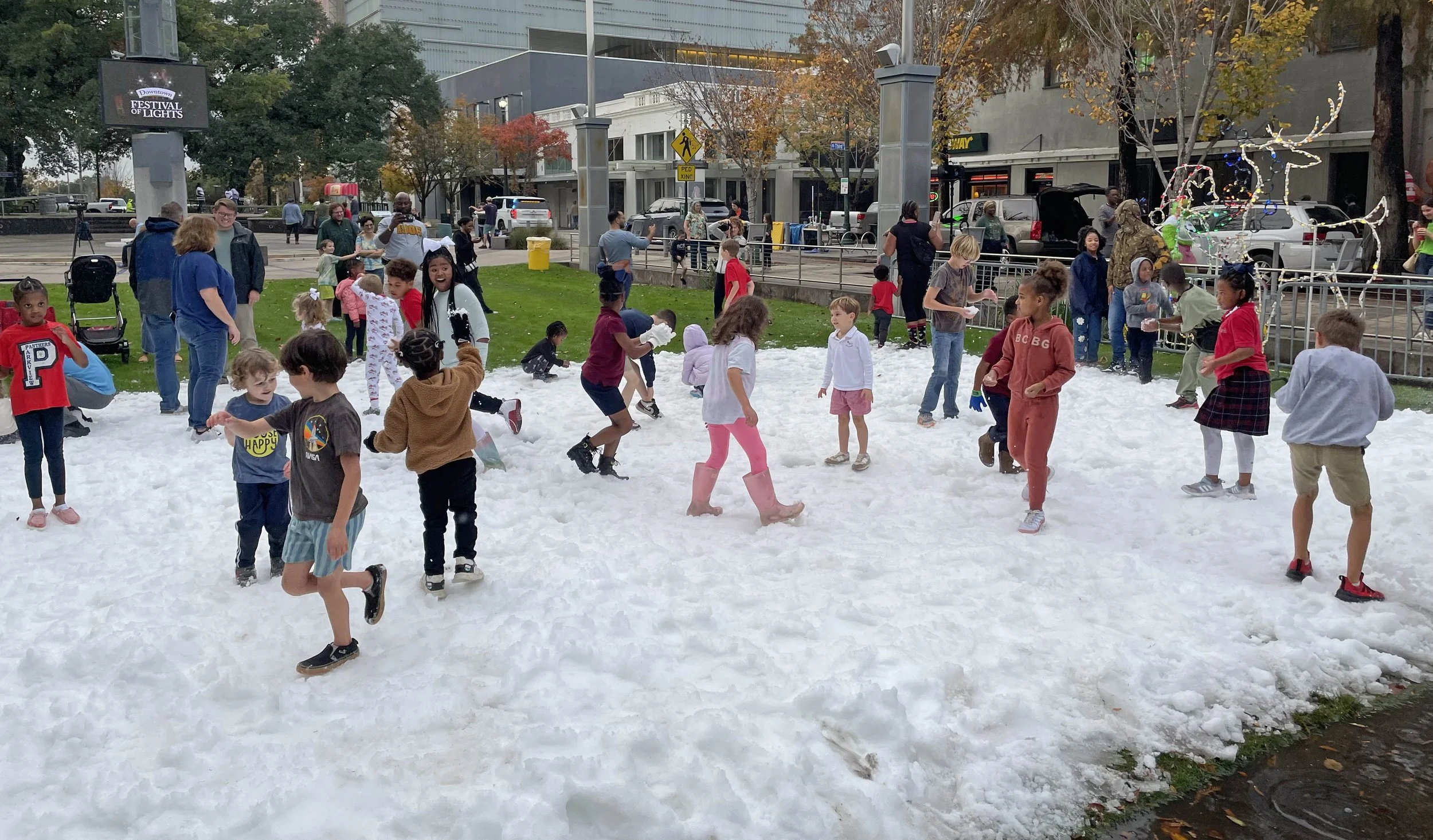 Children and adults playing on a snow-covered area in an urban park during a festival of lights event, with city buildings, trees, and street signs in the background.