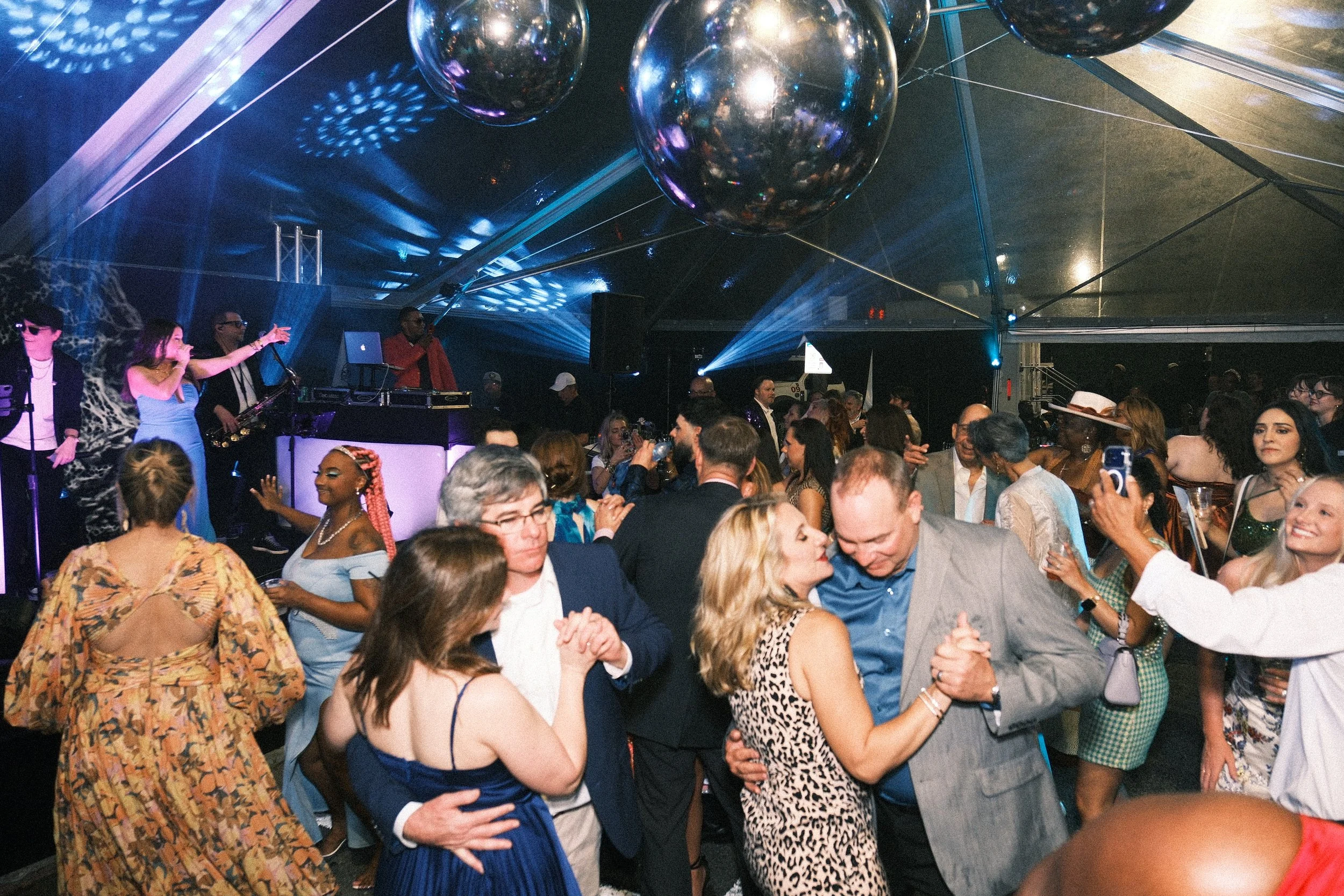 A lively indoor party scene with people dancing and socializing under a tent ceiling decorated with large reflective black balloons and blue lighting, while a DJ plays music.