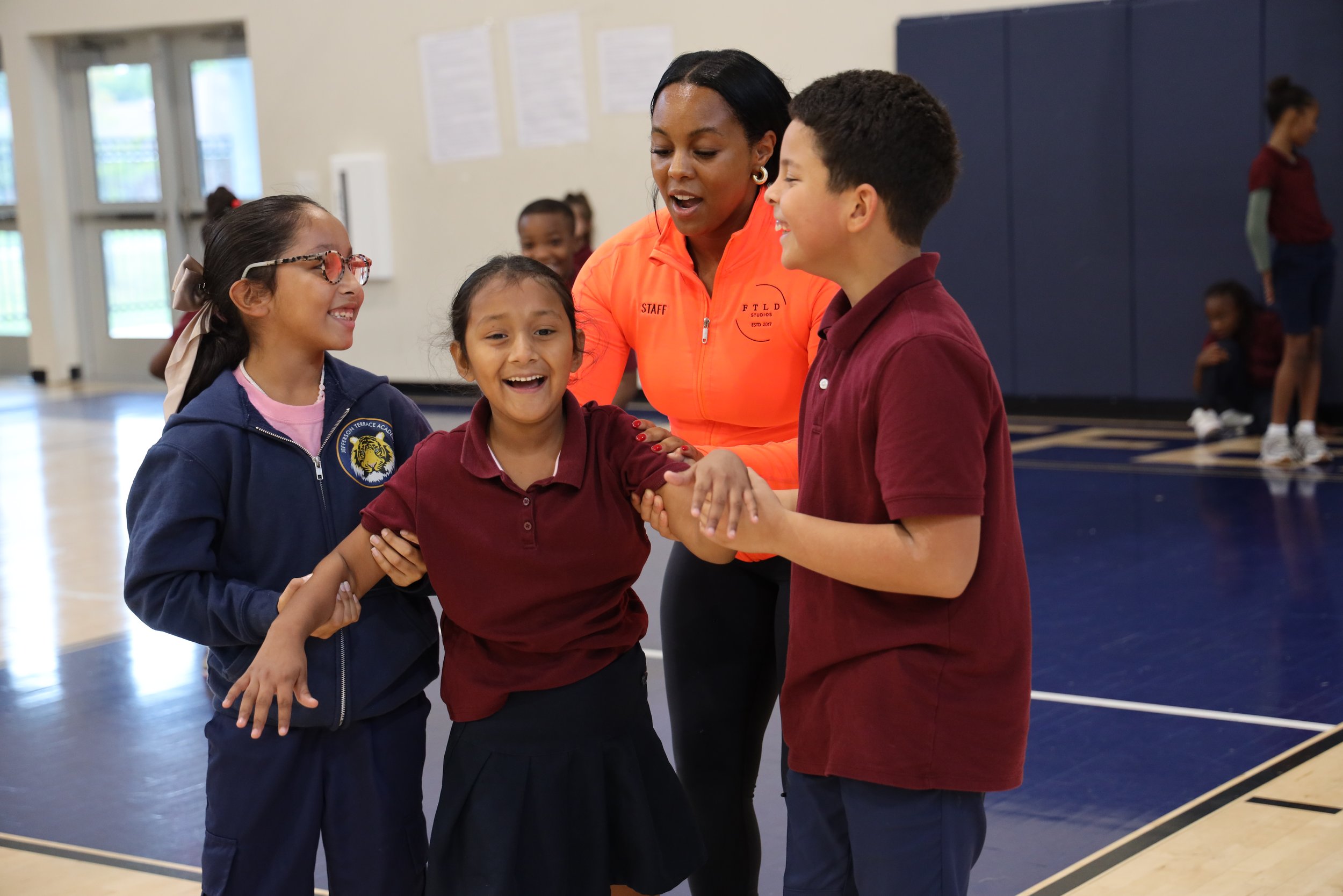 A group of children and a woman are gathered in a gymnasium. The woman, wearing an orange jacket labeled 'STAFF,' is talking to a boy and a girl, smiling and holding their hands. The children are smiling and one girl is wearing glasses. In the background, other children are seen playing or standing.