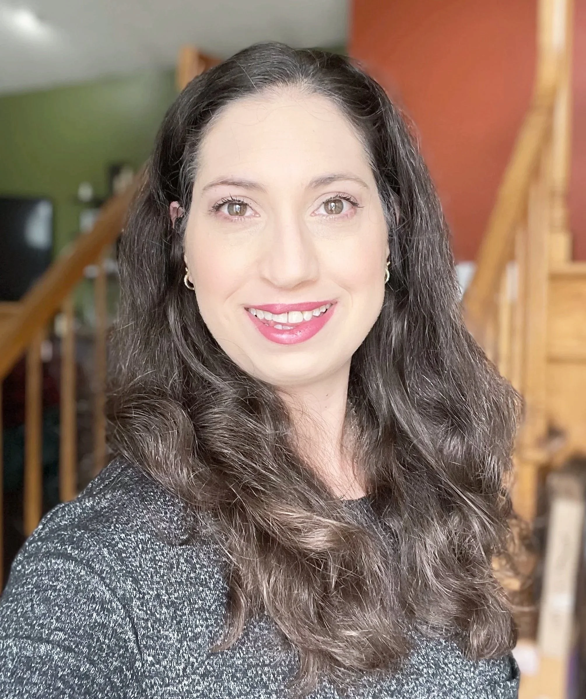 Close-up of a woman with long, wavy brown hair and light makeup, smiling, indoors with wooden staircase in the background.