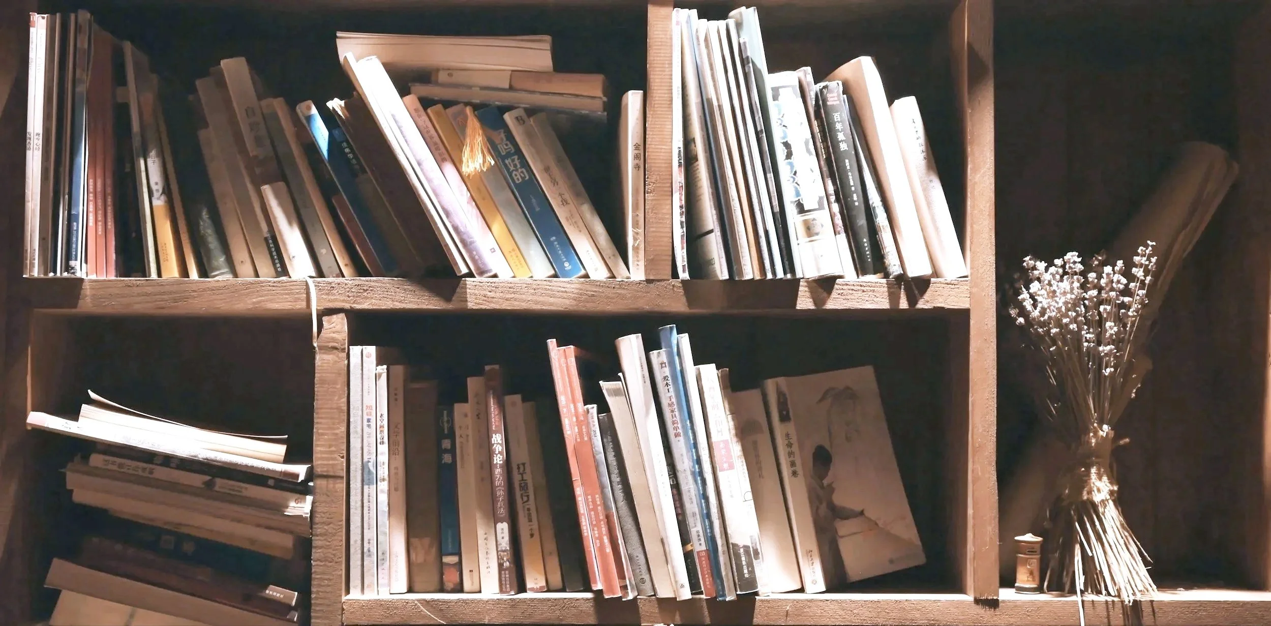 A wooden bookshelf with various books and magazines, a framed picture, a vase of flowers, and a small object on a dark background.