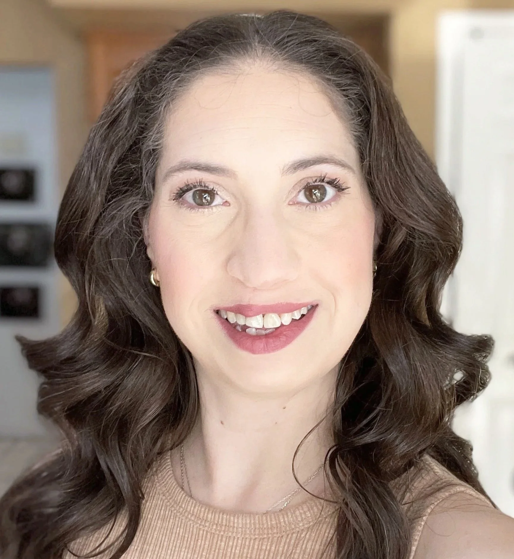 A woman with long wavy brown hair, wearing makeup, a beige top, and a delicate necklace, smiling inside a home.