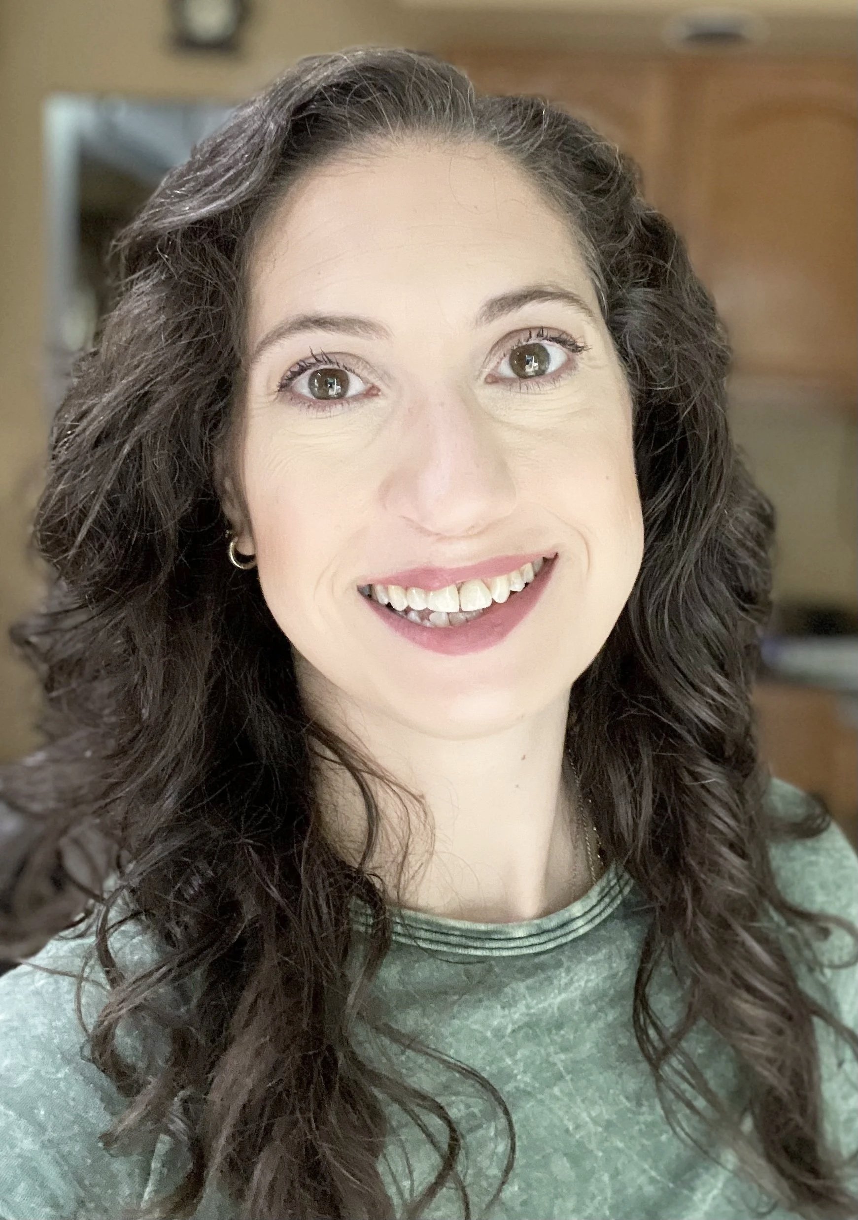 A woman with curly brown hair and hazel eyes smiling, wearing a green top, in a room with wooden cabinets.