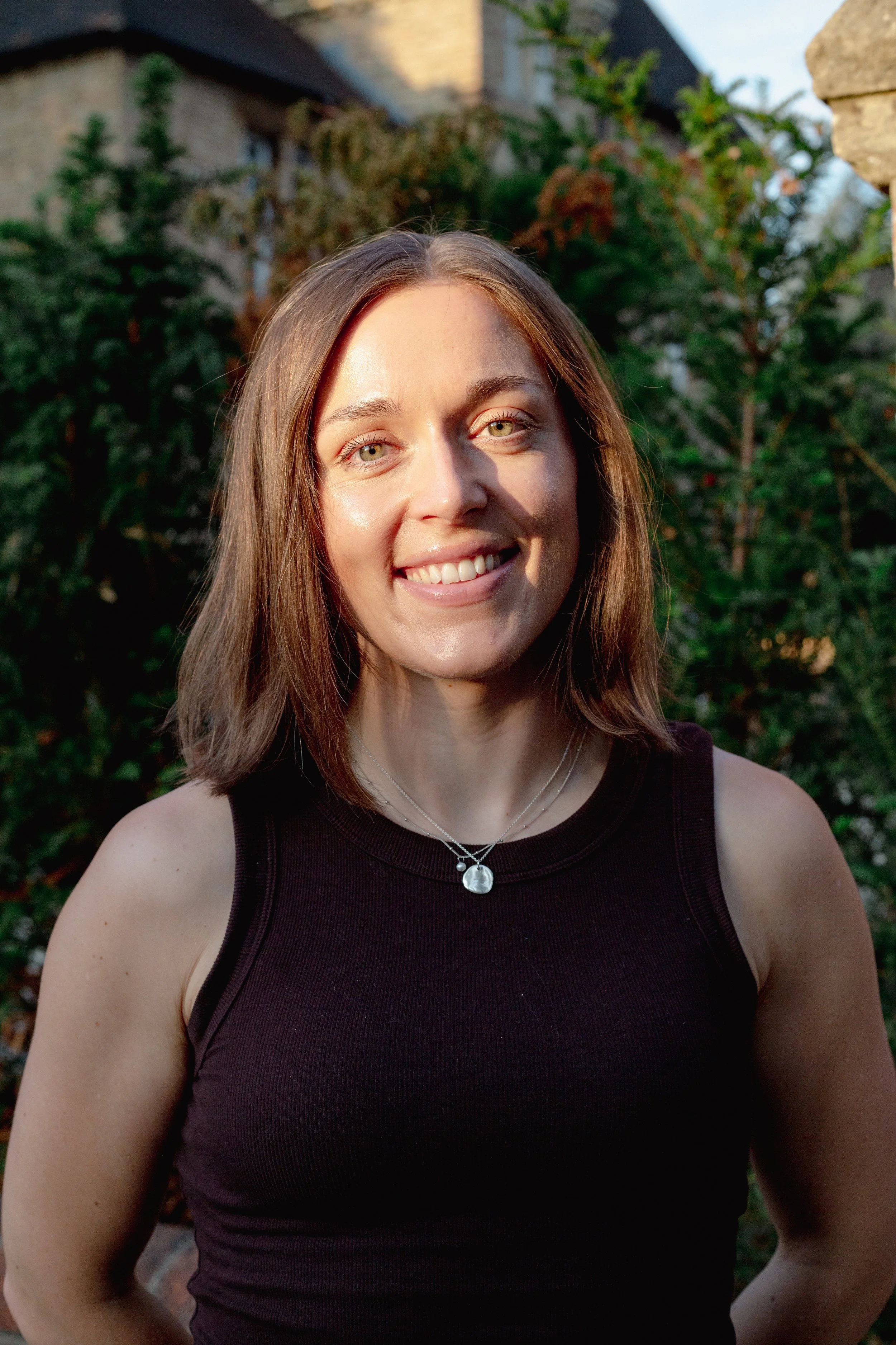 A woman with shoulder-length brown hair and light green eyes smiling outdoors. She is wearing a black sleeveless top and two silver necklaces with pendants. The background includes greenery and a stone wall, with a house partially visible behind the foliage.