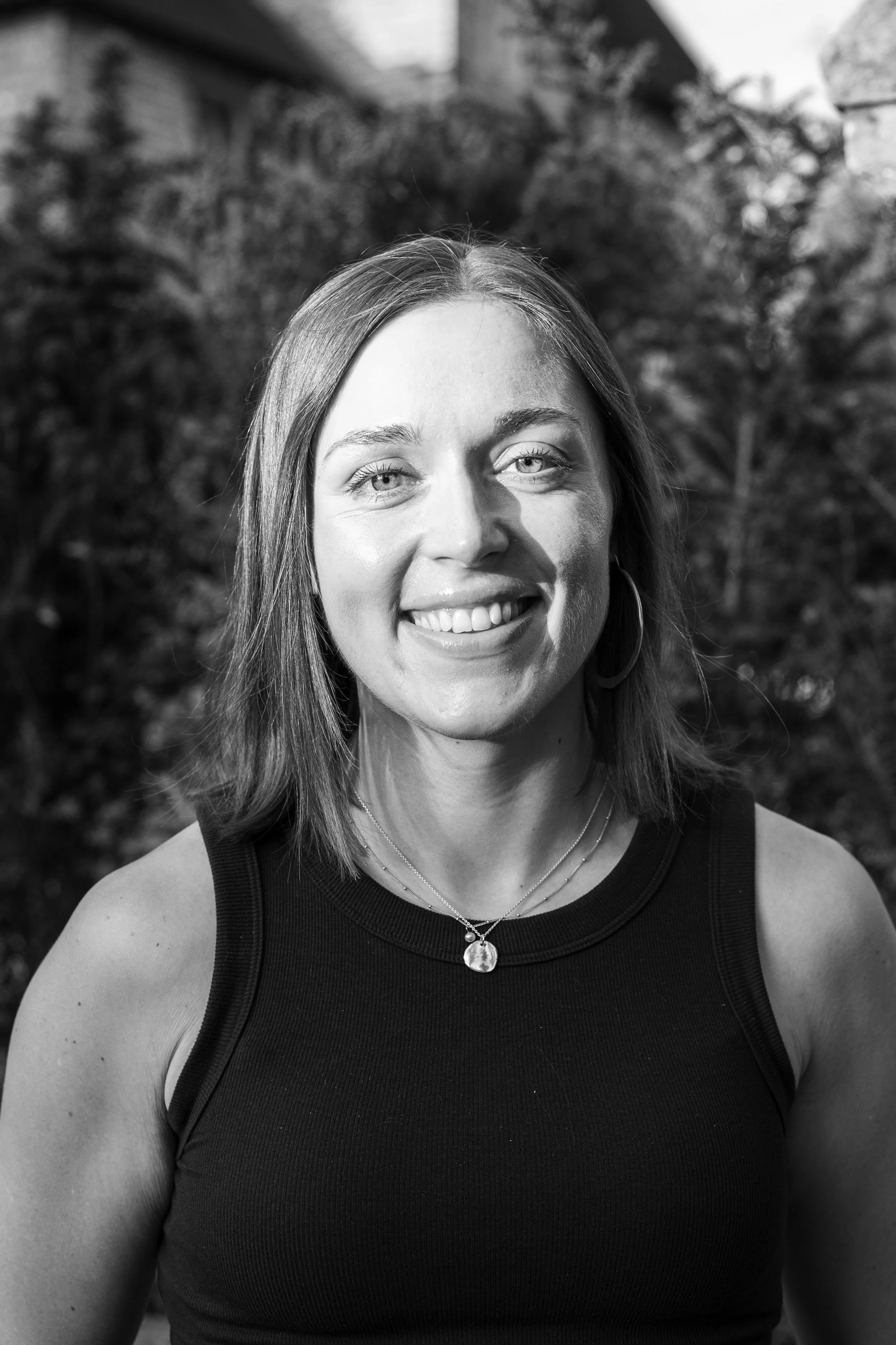 Black and white photo of a smiling woman with long hair, wearing hoop earrings, a necklace, and a sleeveless top, outdoors with blurred foliage in background.