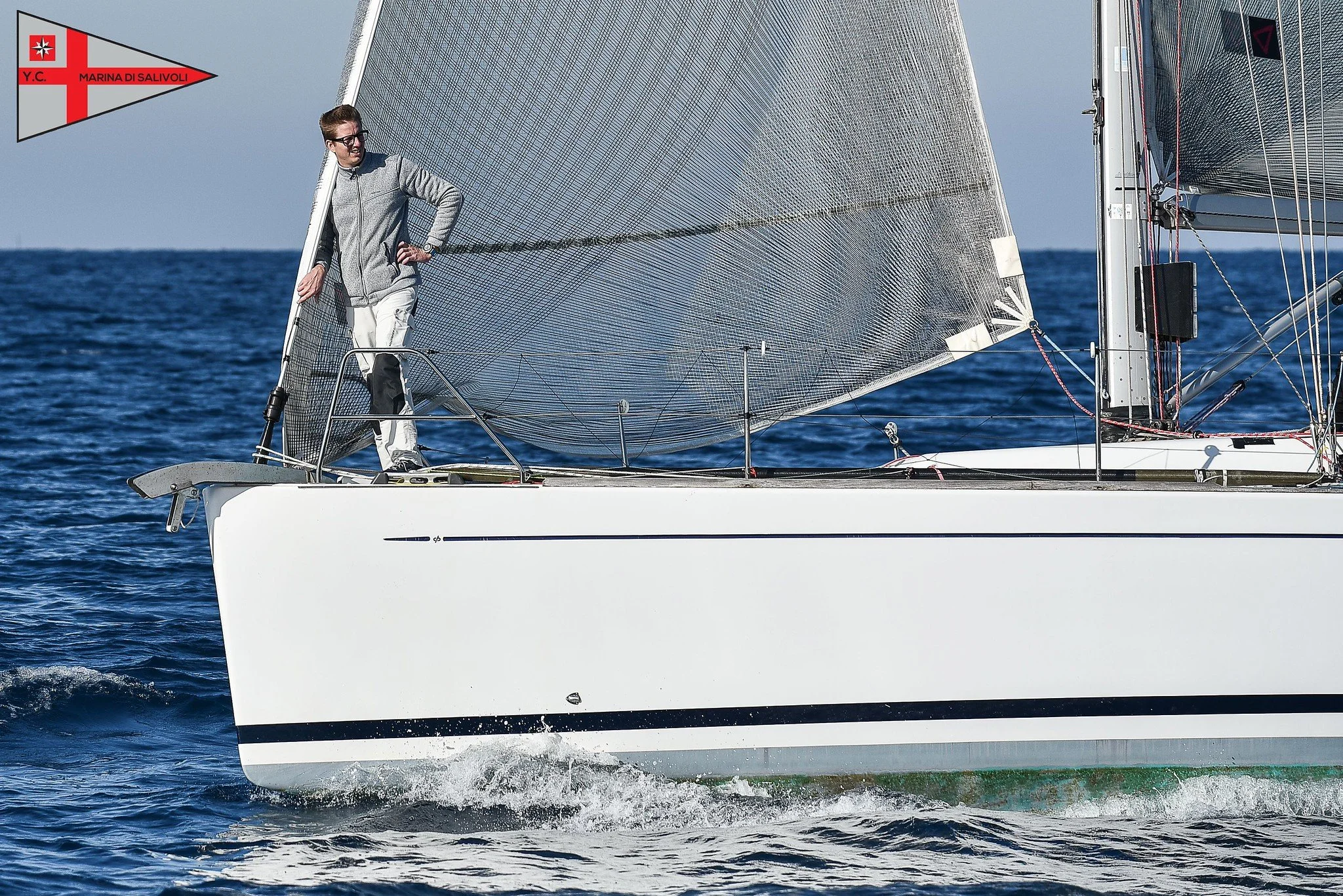 Giovane uomo in piedi sul ponte di una barca a vela bianca con vele al vento, in acqua aperta con mare calmo e cielo sereno.
