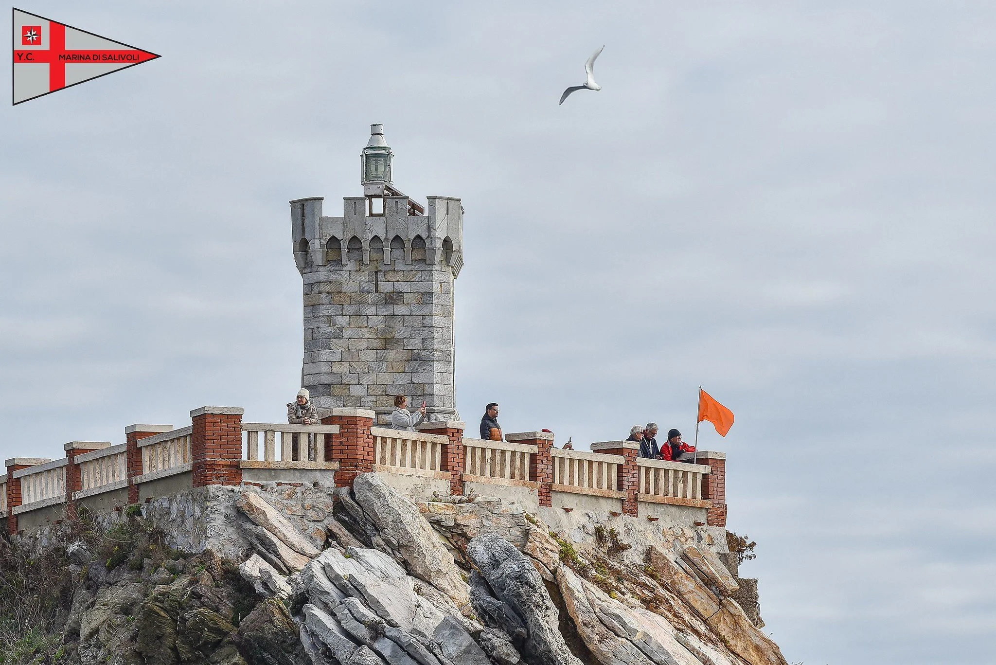 Torri e vista di una scogliera con persone che si godono il panorama, bandiera arancione e un gabbiano in volo, nel sali-livoli di una struttura in pietra con parapetto.