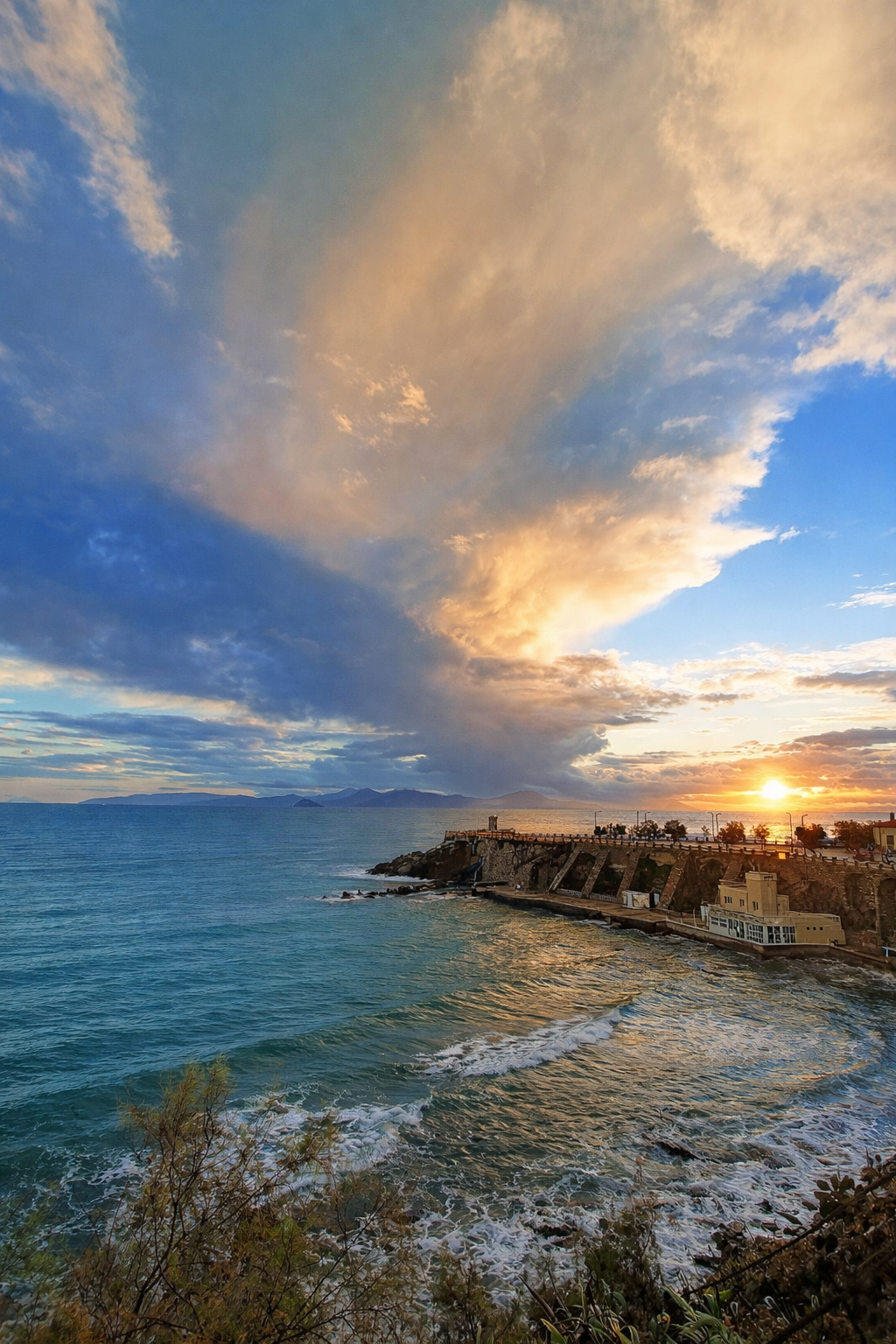 Scorcio del mare con onde che si infrangono sulla spiaggia, un promontorio con edifici e alberi sullo sfondo, e un cielo con nuvole colorate dal tramonto.