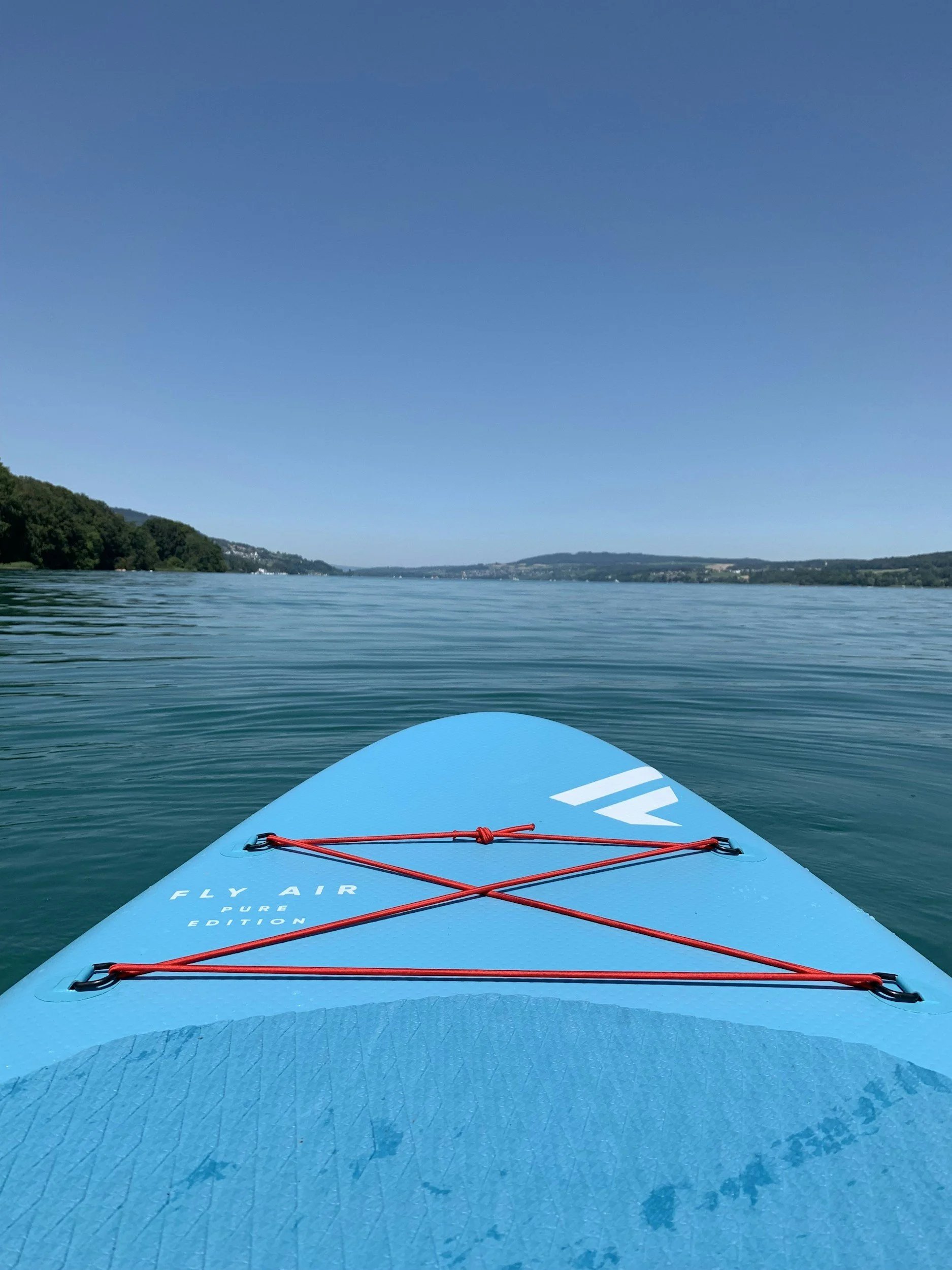 Vista frontale di una tavola da stand-up paddle su un lago calmo, con panorama di colline e cielo sereno.
