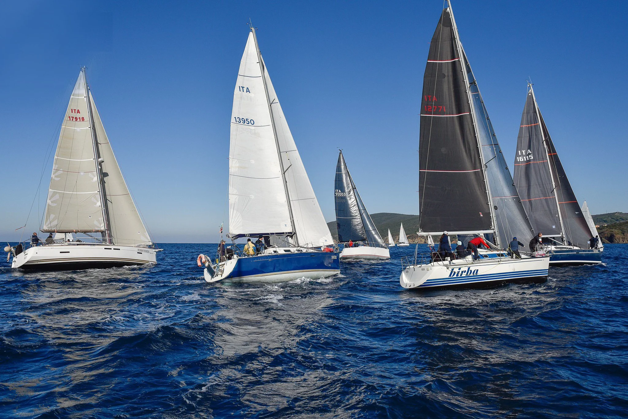 Cinque barche a vela con persone a bordo che navigano in mare con un paesaggio di scogli e colline sullo sfondo, cielo chiaro e sole.