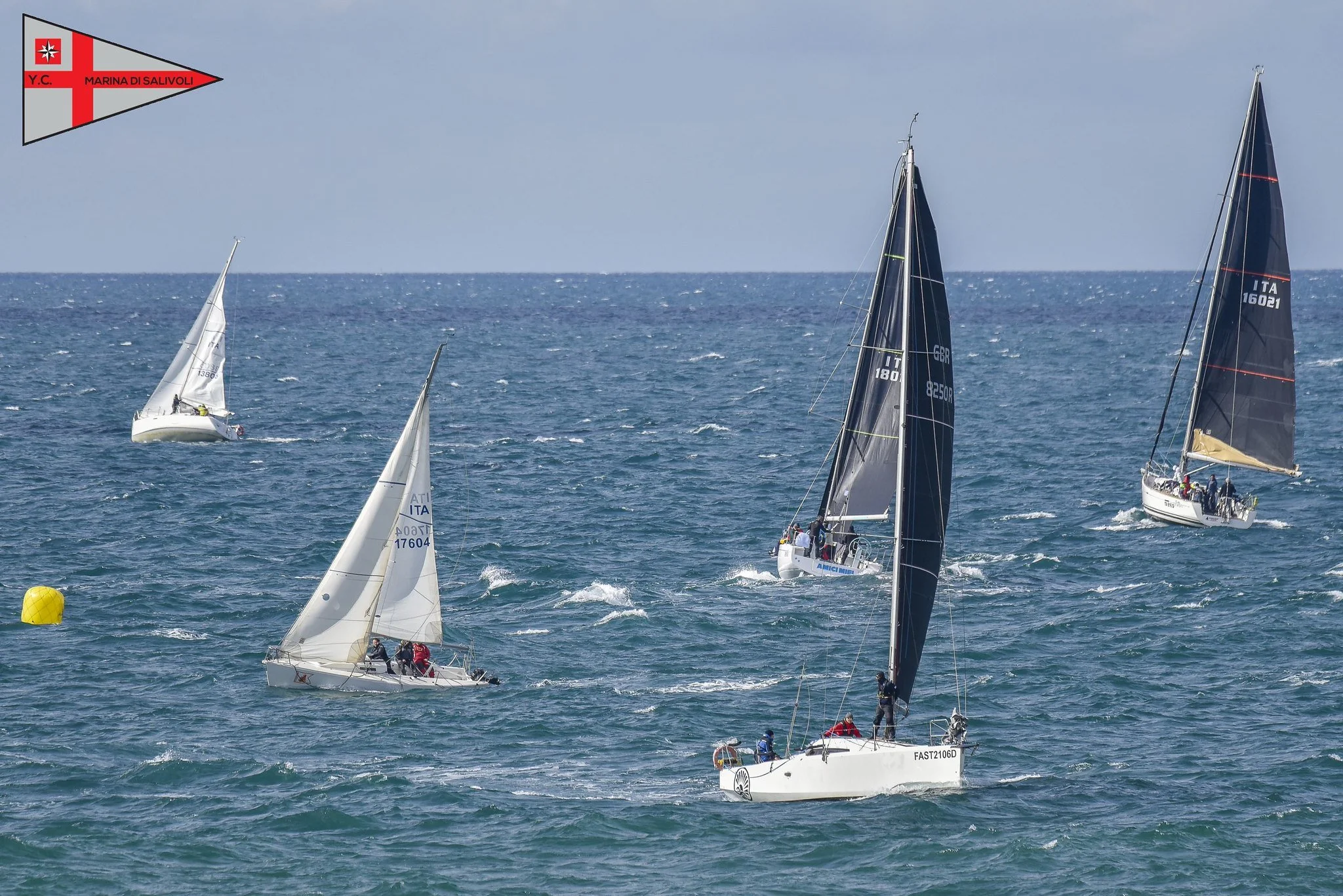 Barche a vela durante una regata in mare, con vela nera, bianca e di altri colori, e boe gialle segnali di percorso.