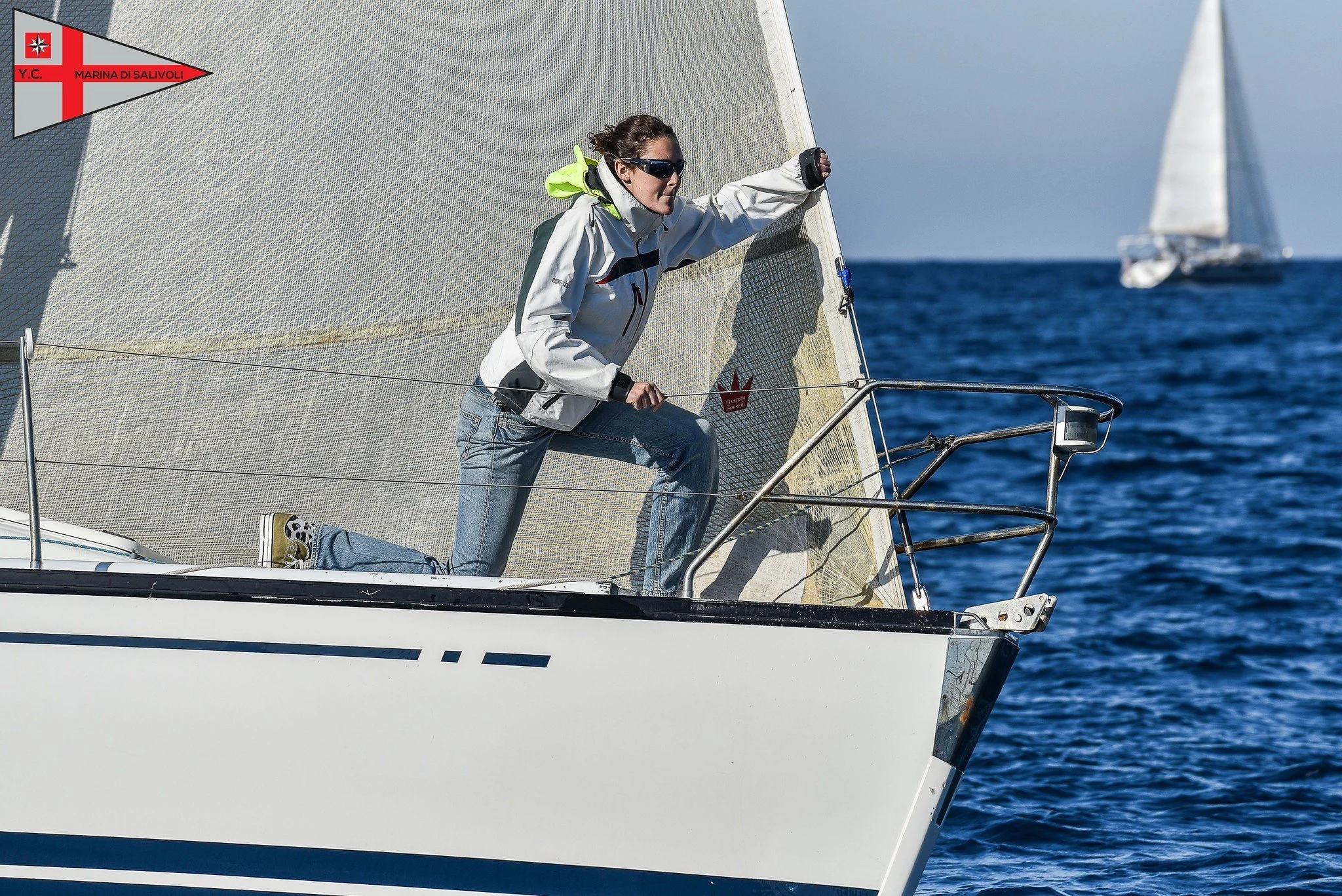 Una donna che naviga su una barca a vela durante il giorno, indossa occhiali da sole e una giacca bianca, con il mare blu sullo sfondo