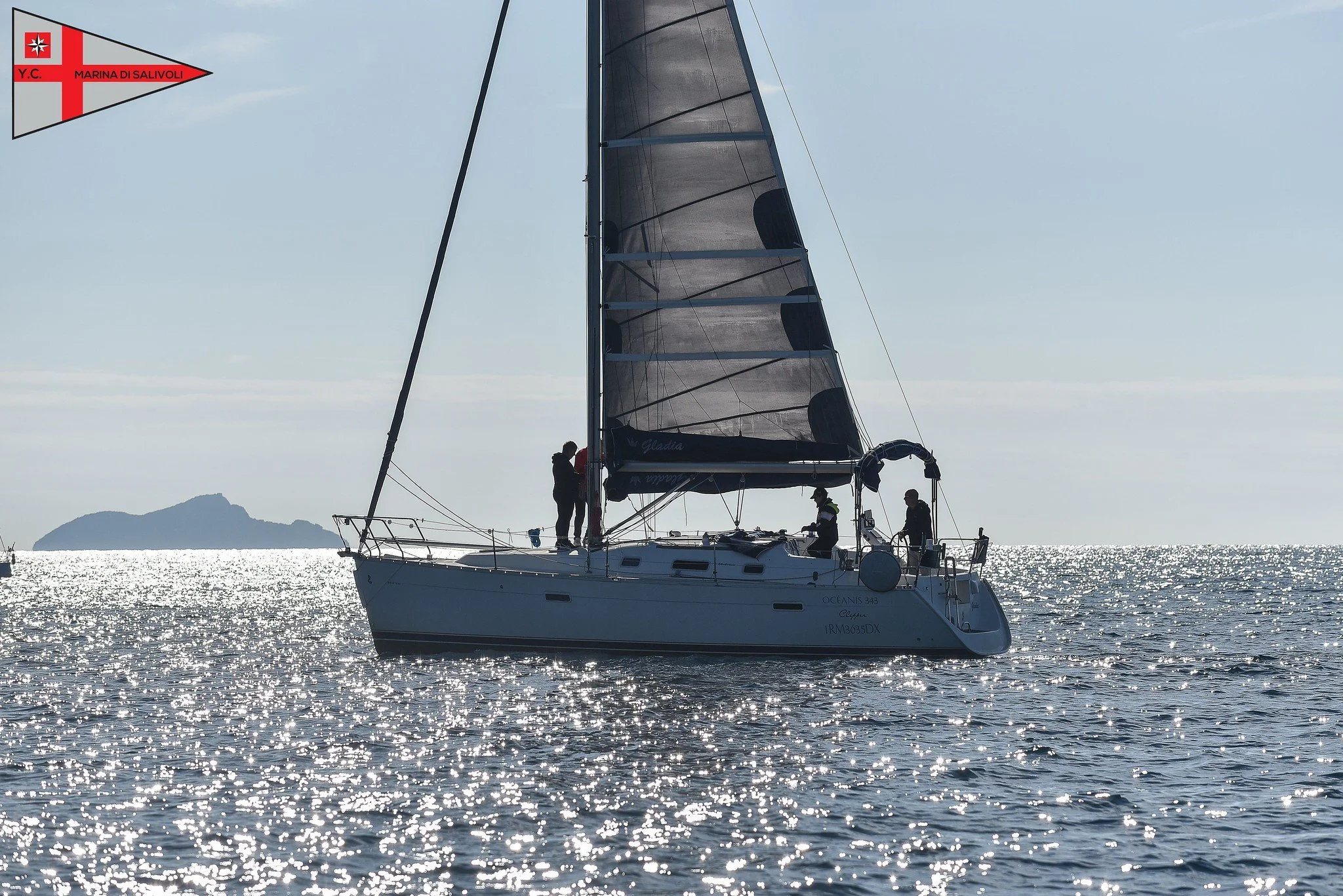 Una barca a vela in mare con quattro persone a bordo, il sole che riflette sull'acqua e una piccola isola sullo sfondo.
