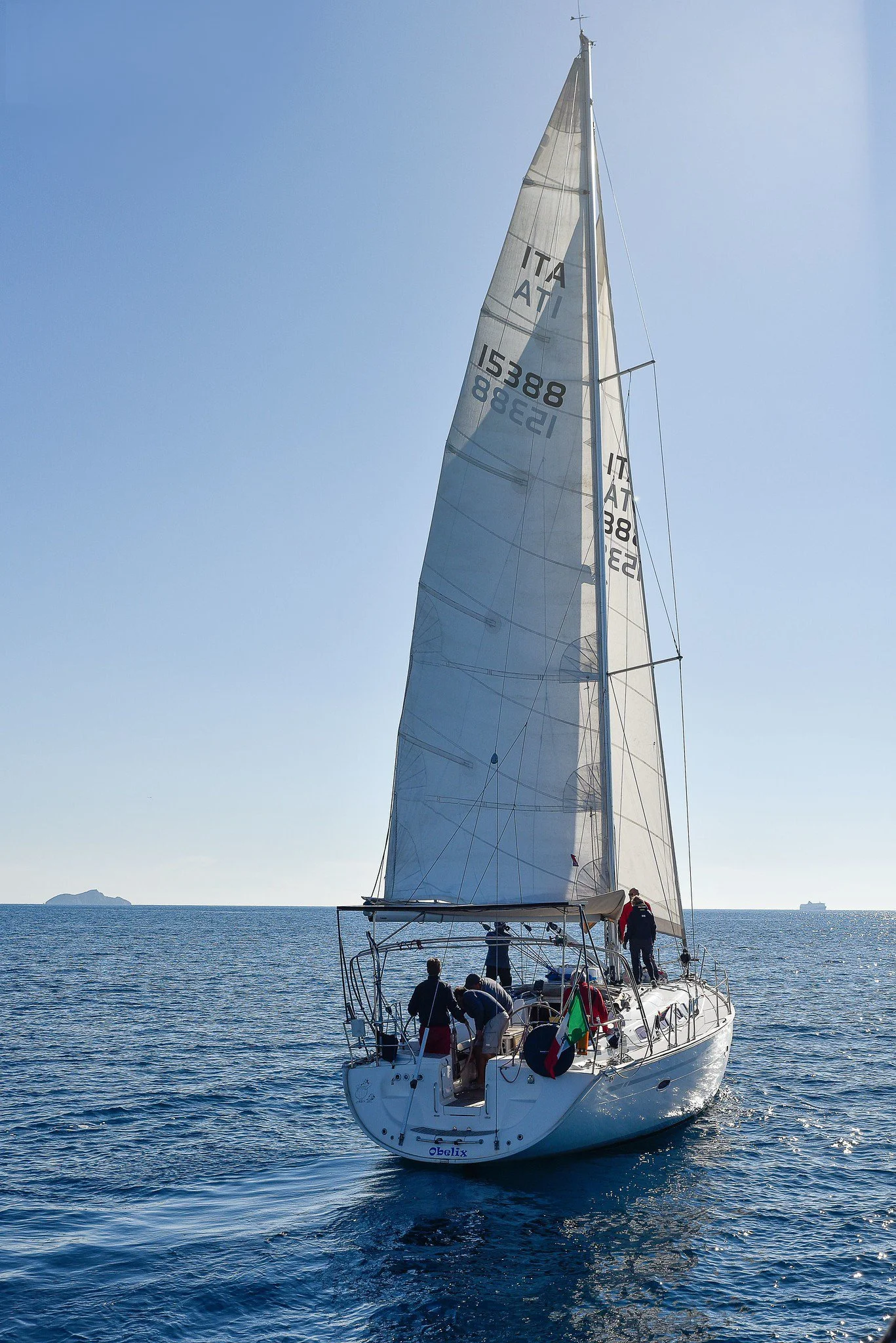 Barca a vela in mare con cielo sereno, con alcune persone a bordo che si preparano a navigare. Sullo sfondo si vede una piccola isola e alcune navi in lontananza.