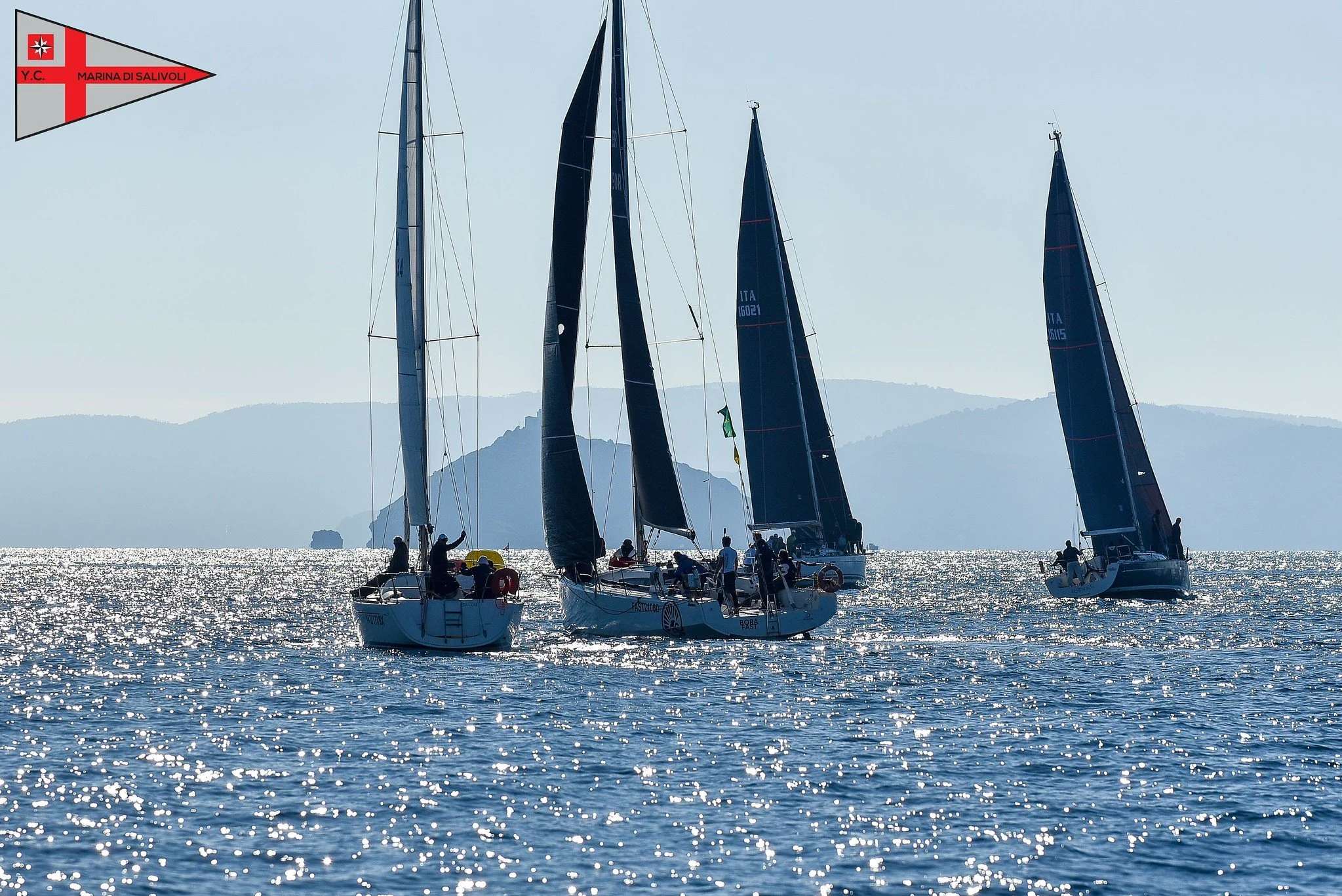 Quattro barche a vela in mare al tramonto, con onde scintillanti e montagne sullo sfondo.