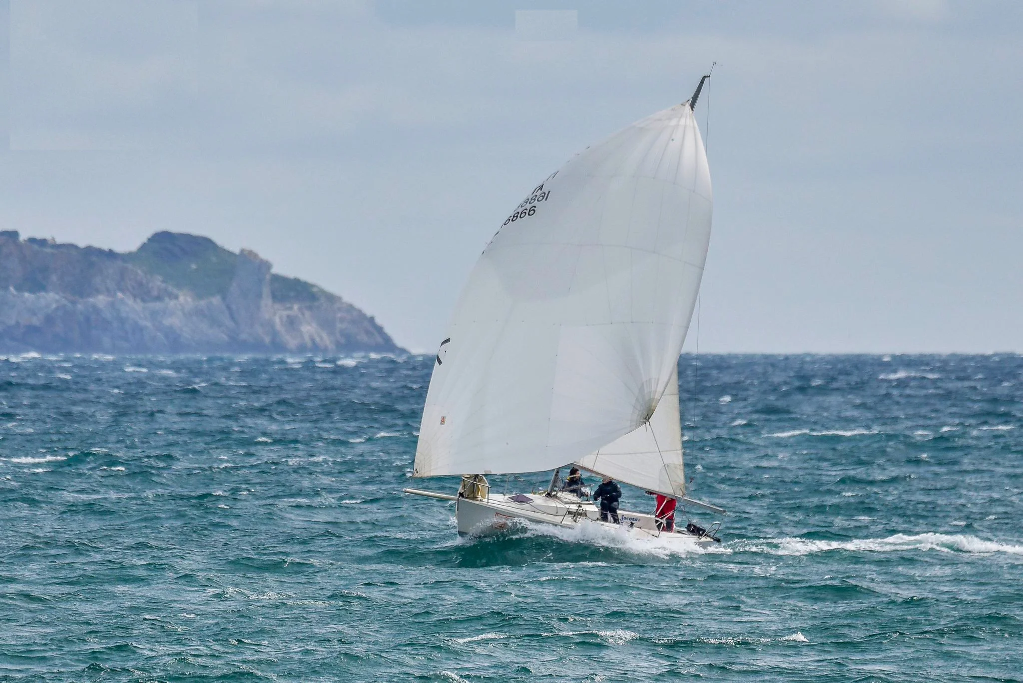 Una barca a vela in mare con un'alta vela bianca, nuvole nel cielo e una costa rocciosa sullo sfondo.