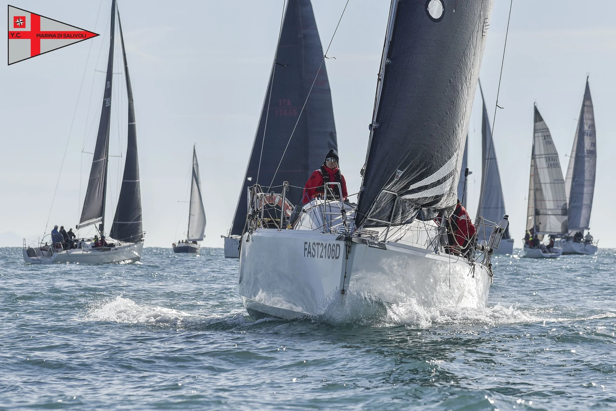 Sailing race with multiple sailboats on the water, sailors dressed in red and black, with sails catching the wind, under a partly cloudy sky.