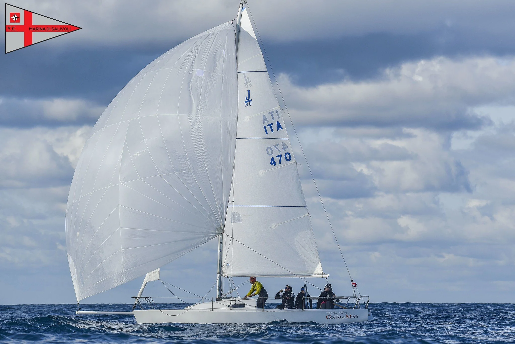 Una barca a vela che naviga sul mare sotto un cielo nuvoloso, con quattro persone a bordo e un grande fiocco bianco che cattura il vento.