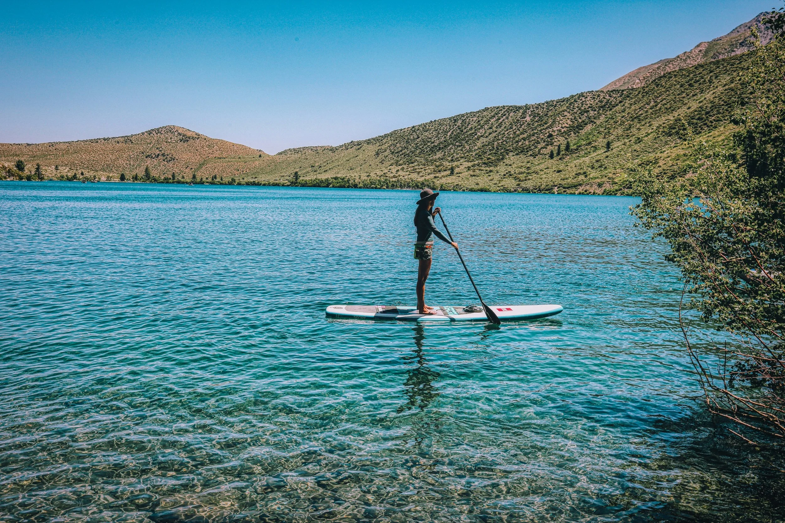 Una donna in costume da bagno e cappello sta pedalando su una tavola a paddle in un lago circondato da colline verdi con cielo sereno.