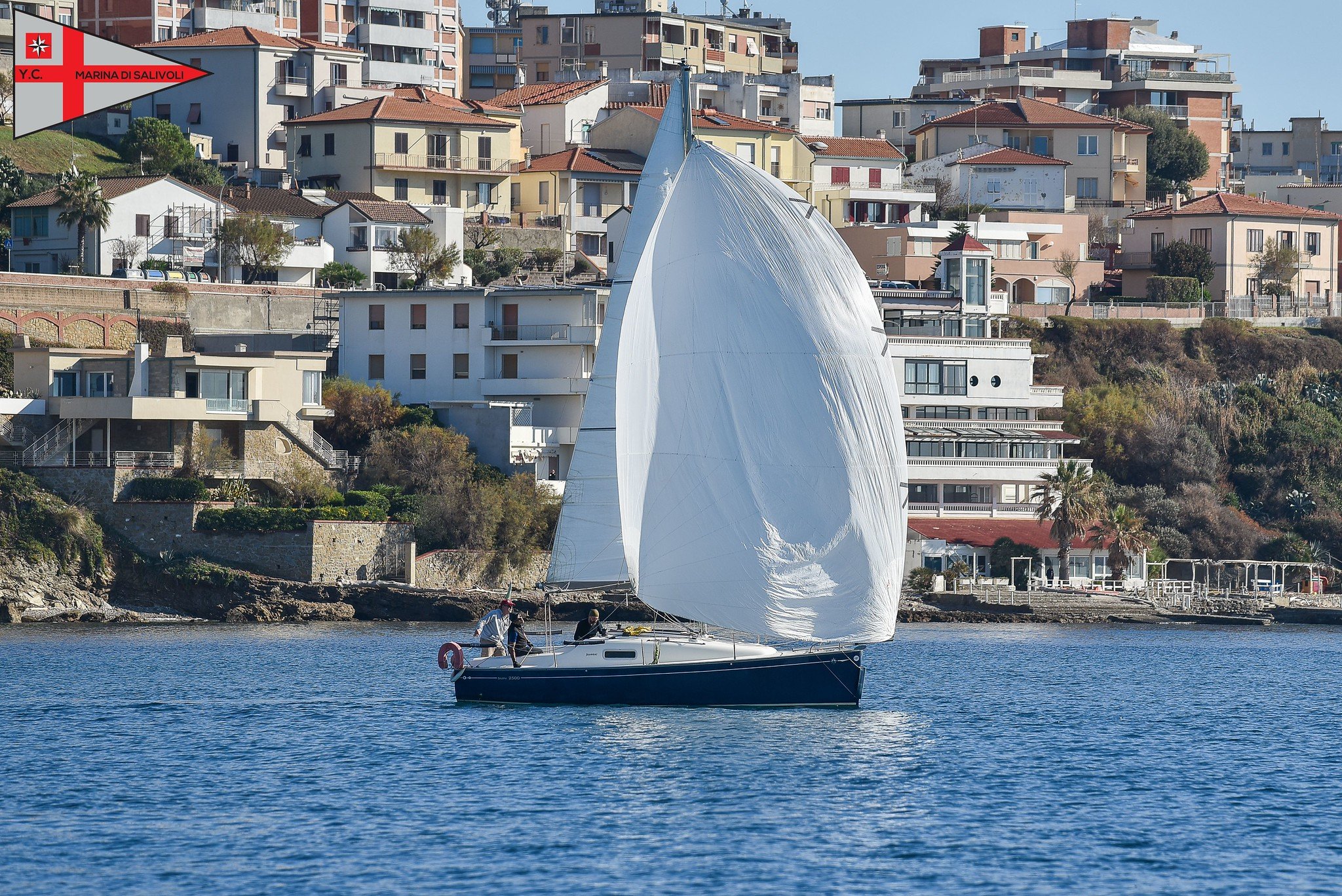 Una barca a vela con vele bianche naviga in mare vicino a una costa abitata con case e edifici di diversi stili e colori, sullo sfondo. Il cielo è sereno.