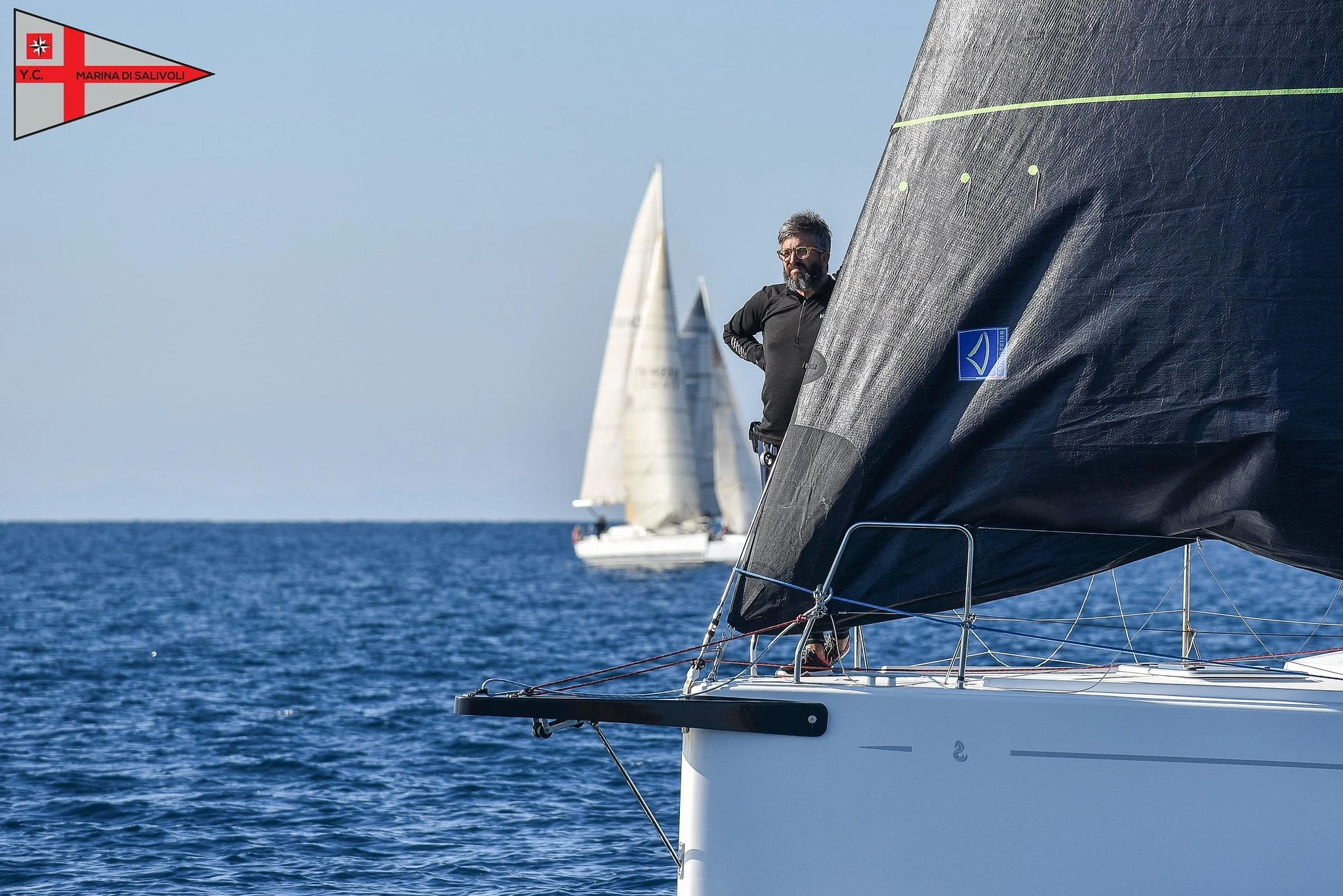 Uomo con barba indossa giacca nera e occhiali da sole a prua di una barca a vela, con altre barche a vela visibili in lontananza sull'acqua.