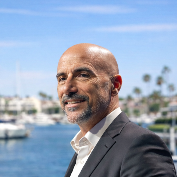 Uomo anziano con barba e testa rasata, vestito con abito da sera, che sorride sullo sfondo di un porto con barche, palme e un cielo blu.