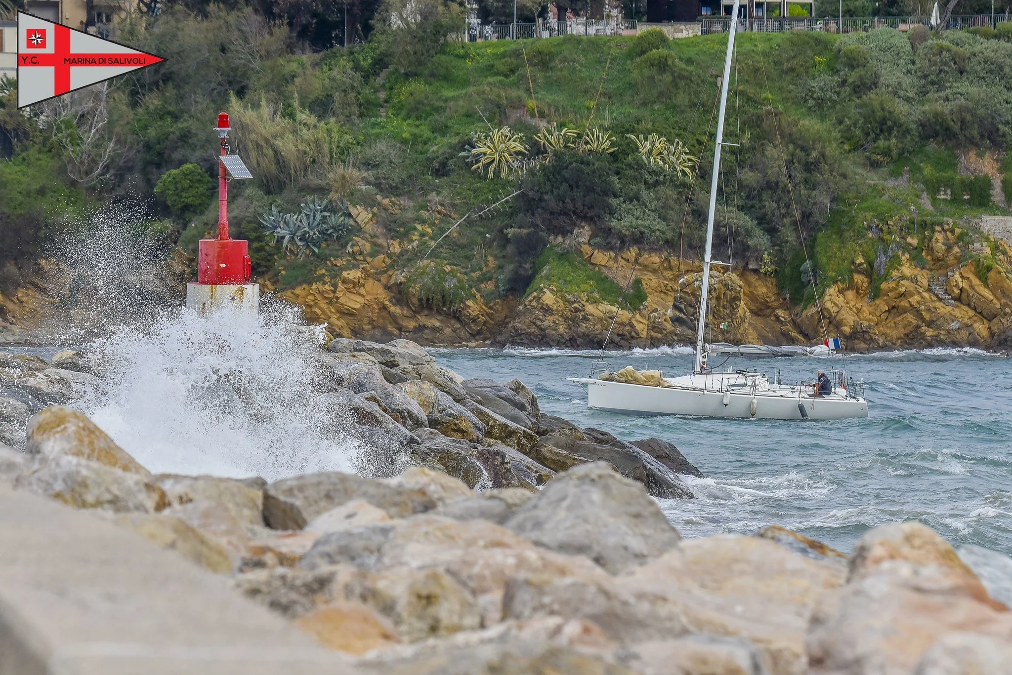 Una barca a vela bianca in mare vicino a un scogliera con vegetazione, con un faro rosso e bianco sulla riva rocciosa e onde che si infrangono contro le rocce.