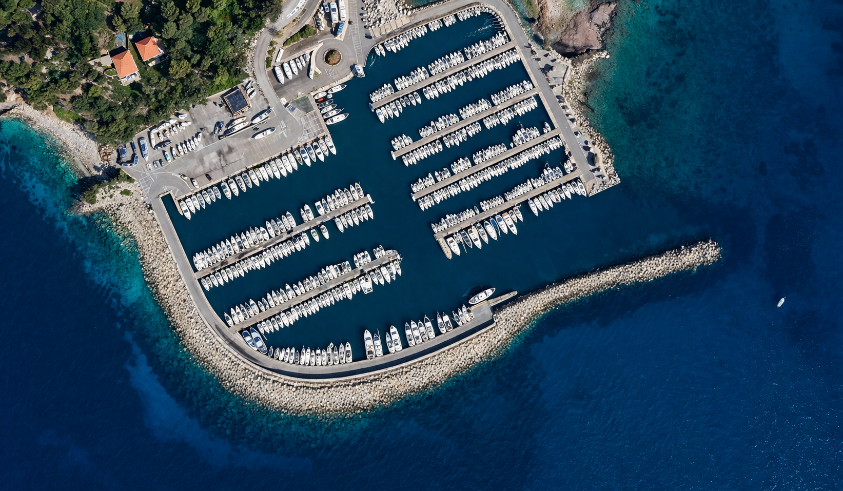 Veduta aerea di un molo con barche ormeggiate, delimitato da pareti di pietra, con area parcheggio e alcuni edifici vicini, sul mare blu.