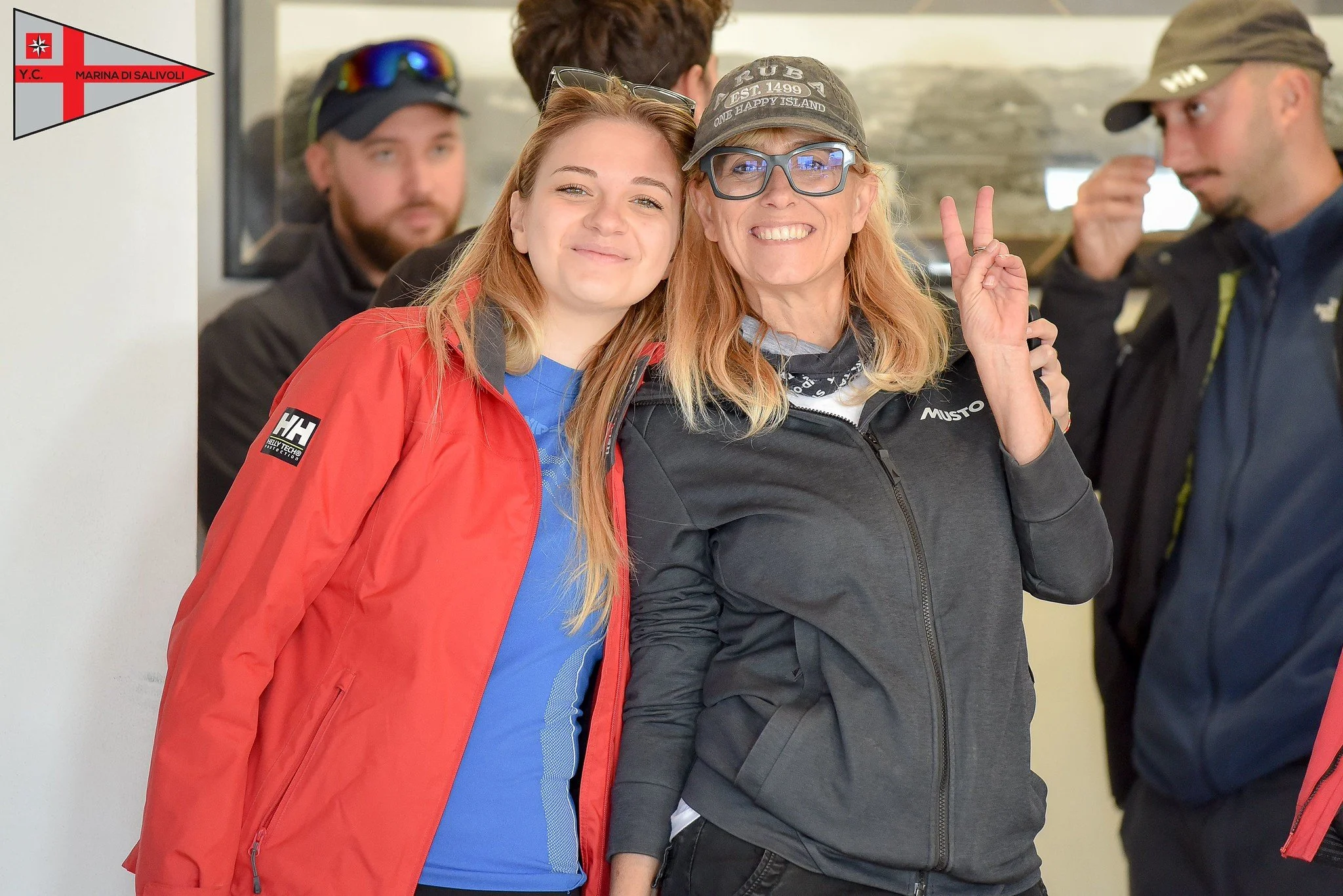 Gruppo di quattro persone sorridenti, due donne in primo piano, una con giacca rossa e l'altra con giacca grigia, entrambe con cappellini e occhiali, che fanno un gesto di pace con le dita. Sfondo di persone in abiti da outdoor, in un ambiente intern
