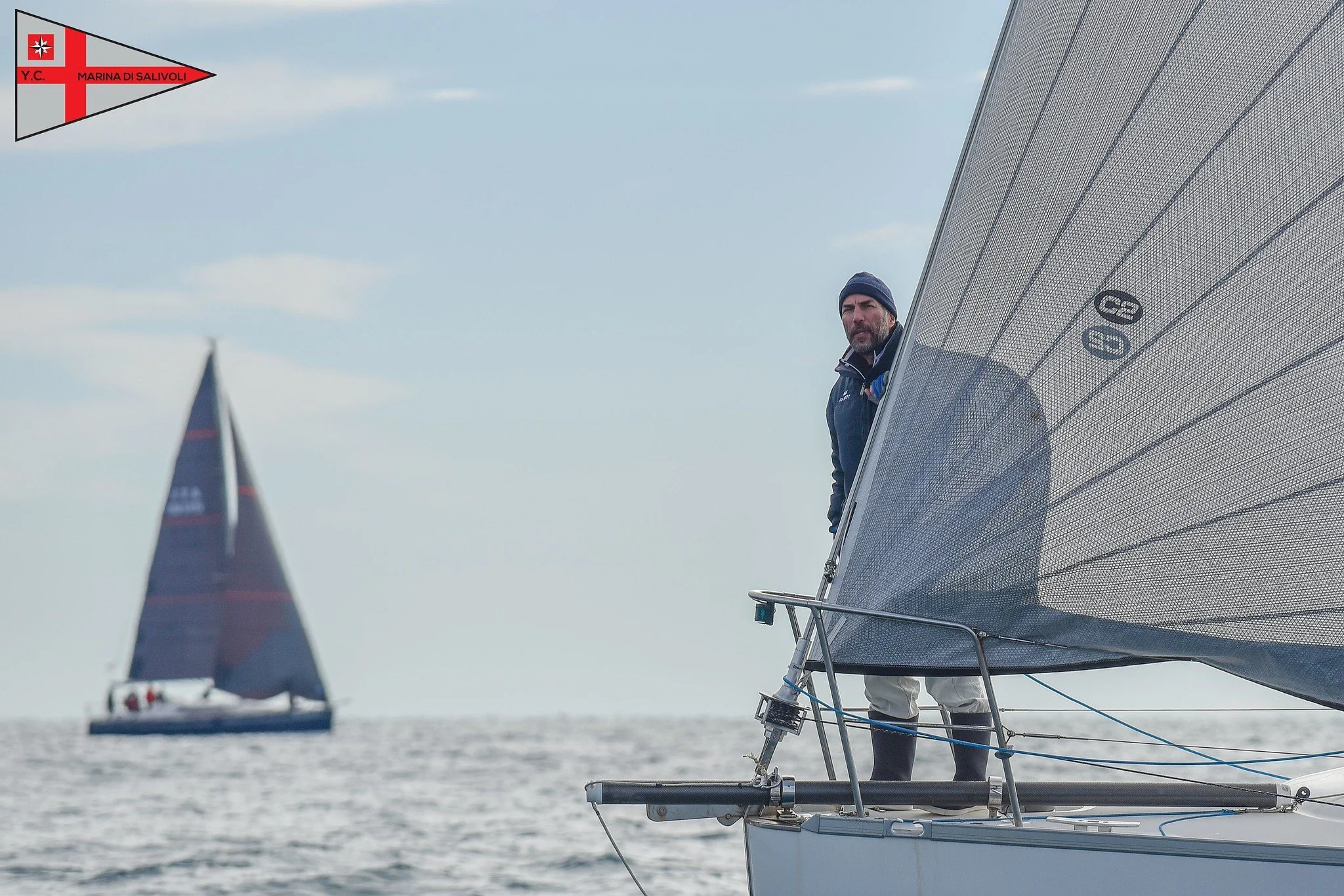 Uomo con giacca e cappello a bordo di una barca a vela, che si affaccia dal fianco, con un altro yacht sullo sfondo in mare, sotto un cielo leggermente nuvoloso.