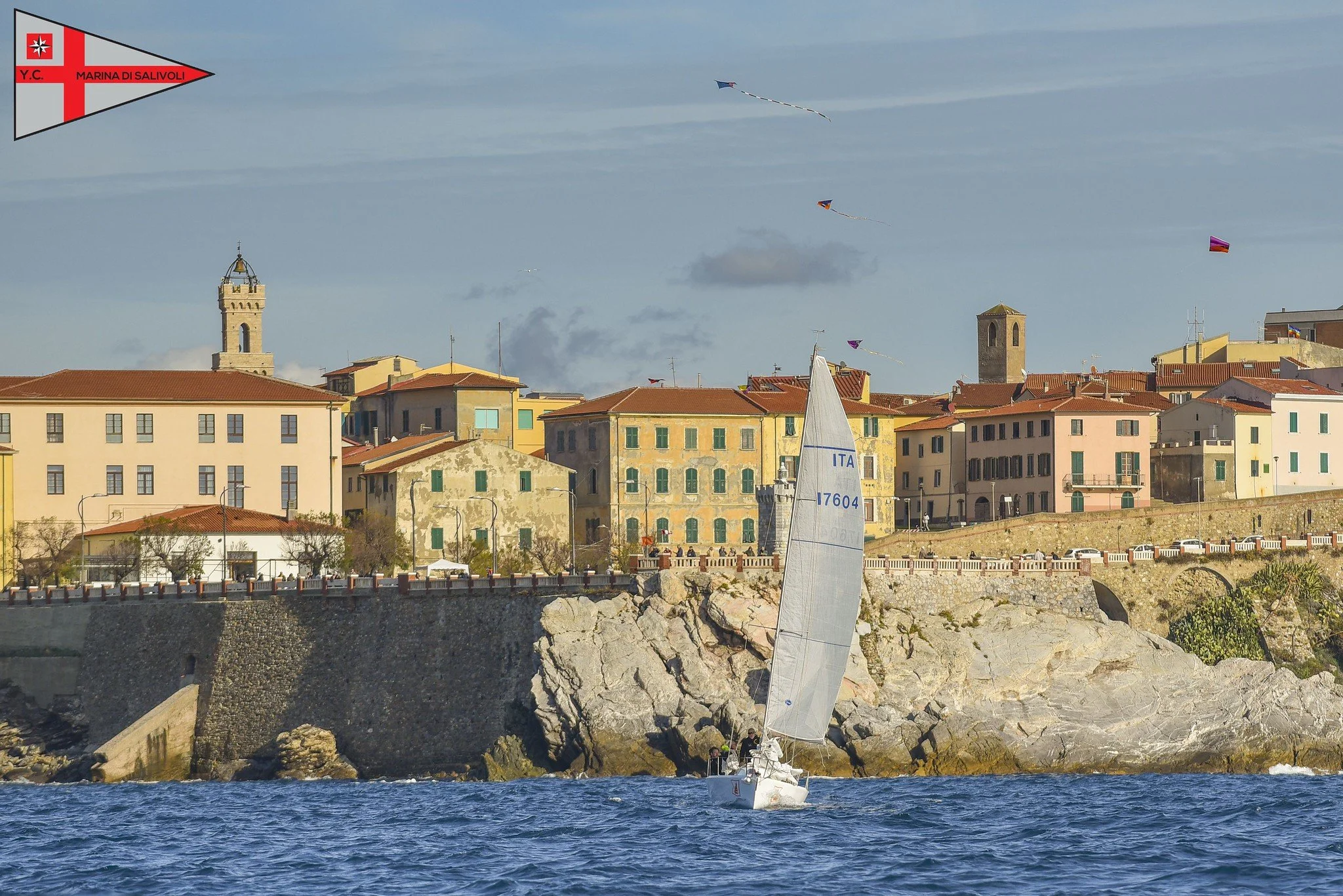 Veduta di un paesaggio costiero con edifici colorati e un porto, con una barca a vela che naviga in mare.