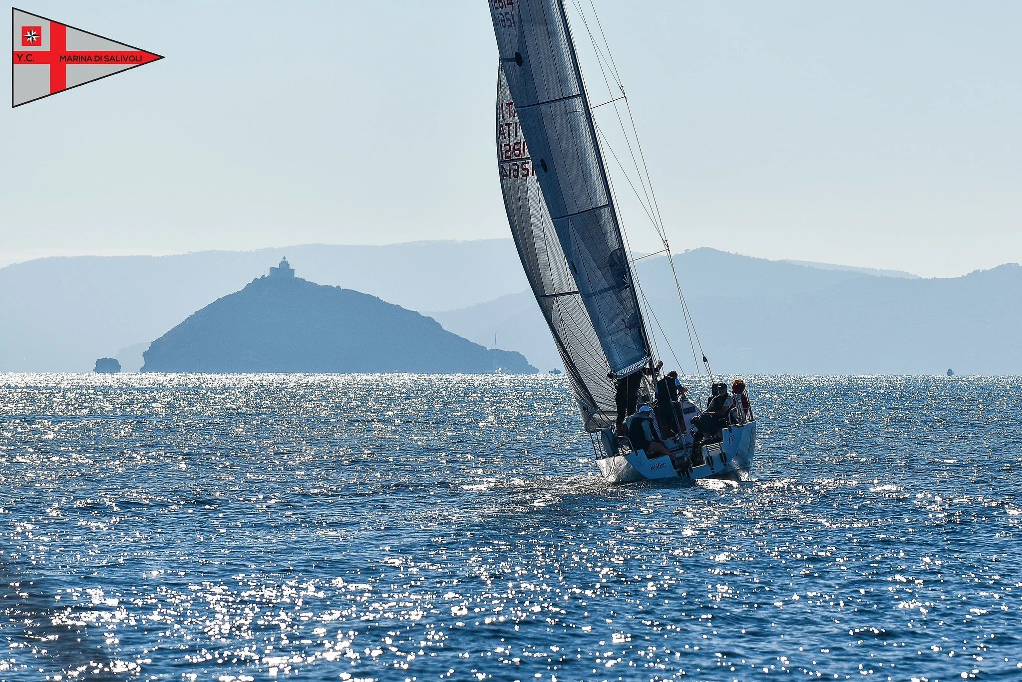 Una barca a vela che naviga in mare con un'isola sullo sfondo e un cielo chiaro.