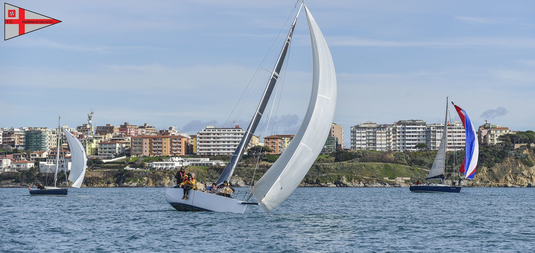 Tre barche a vela che navigano in una baia con edifici residenziali sullo sfondo, sotto un cielo leggermente nuvoloso.