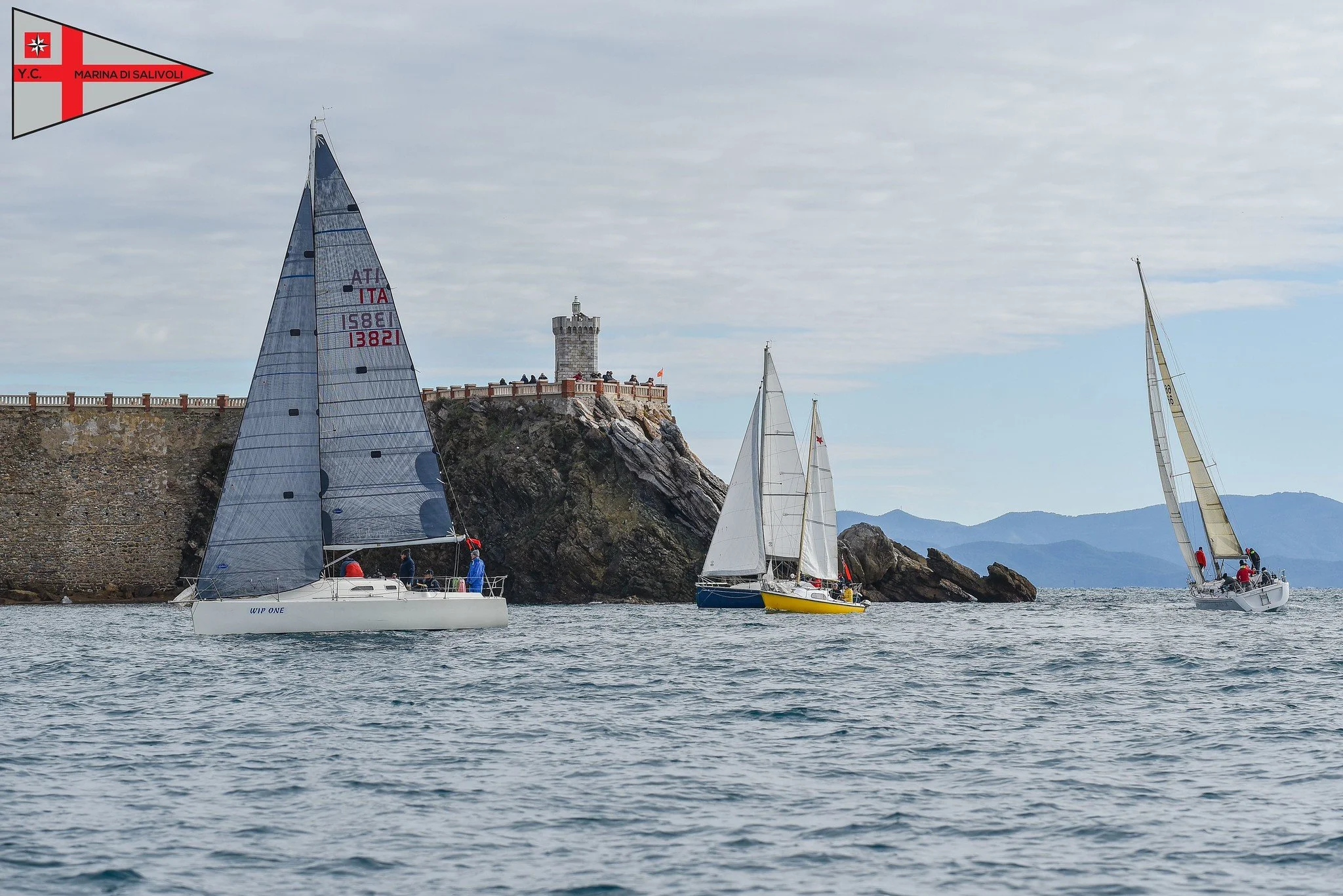 Tre barche a vela navigano vicino a una scogliera con un faro sulla cima, sul mare sotto un cielo nuvoloso.