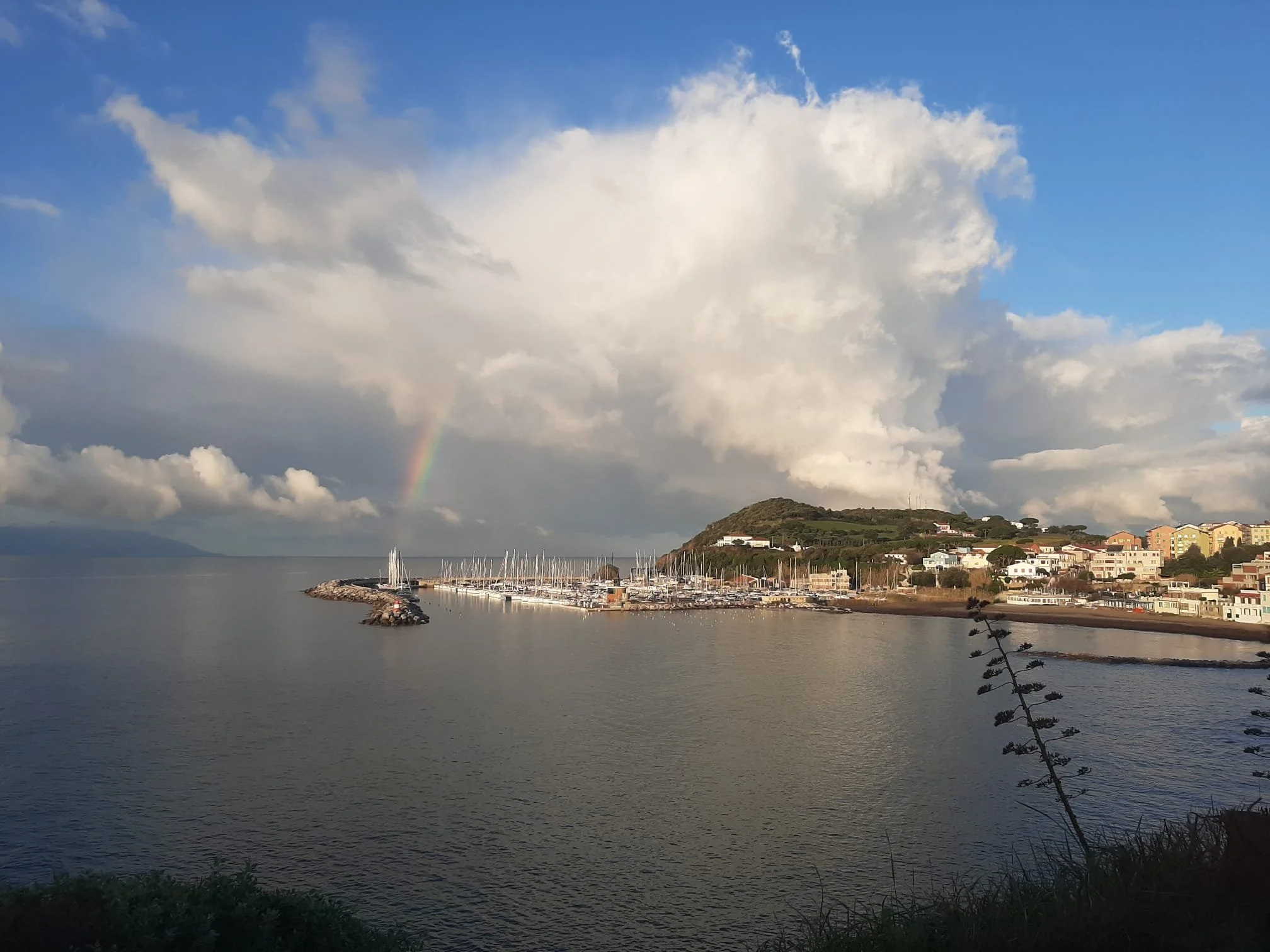 Vasto mare con porto, barche e una collina sullo sfondo, cielo nuvoloso con arcobaleno