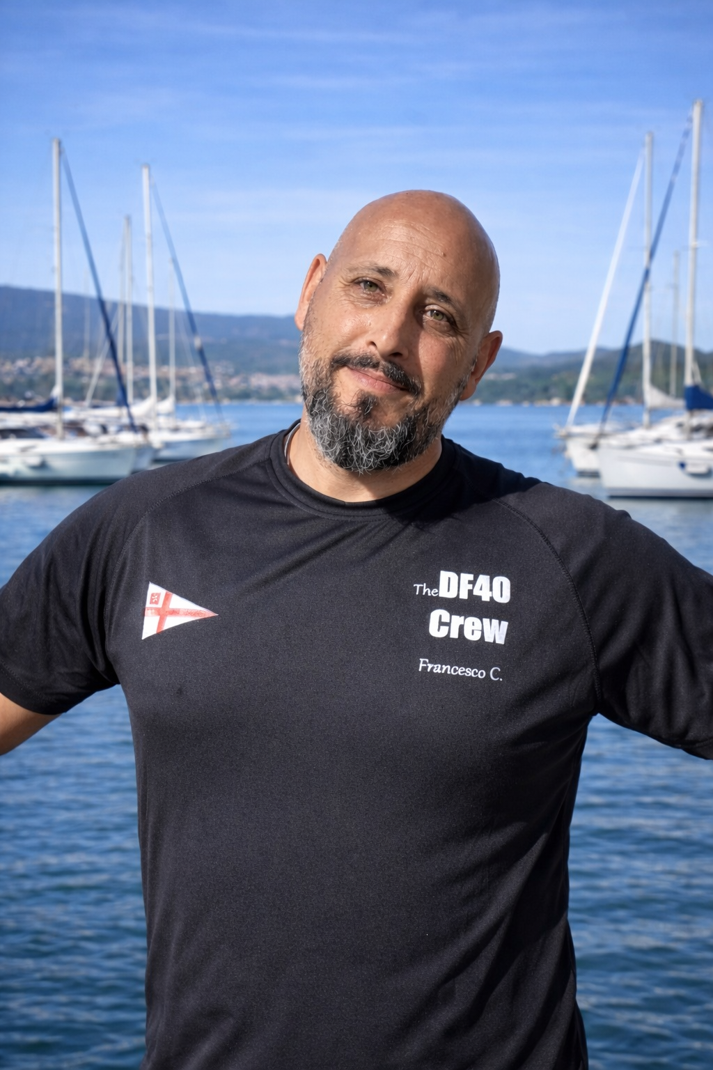 Uomo con barba, maglietta nera con scritte, sfondo di barche a vela nel mare.