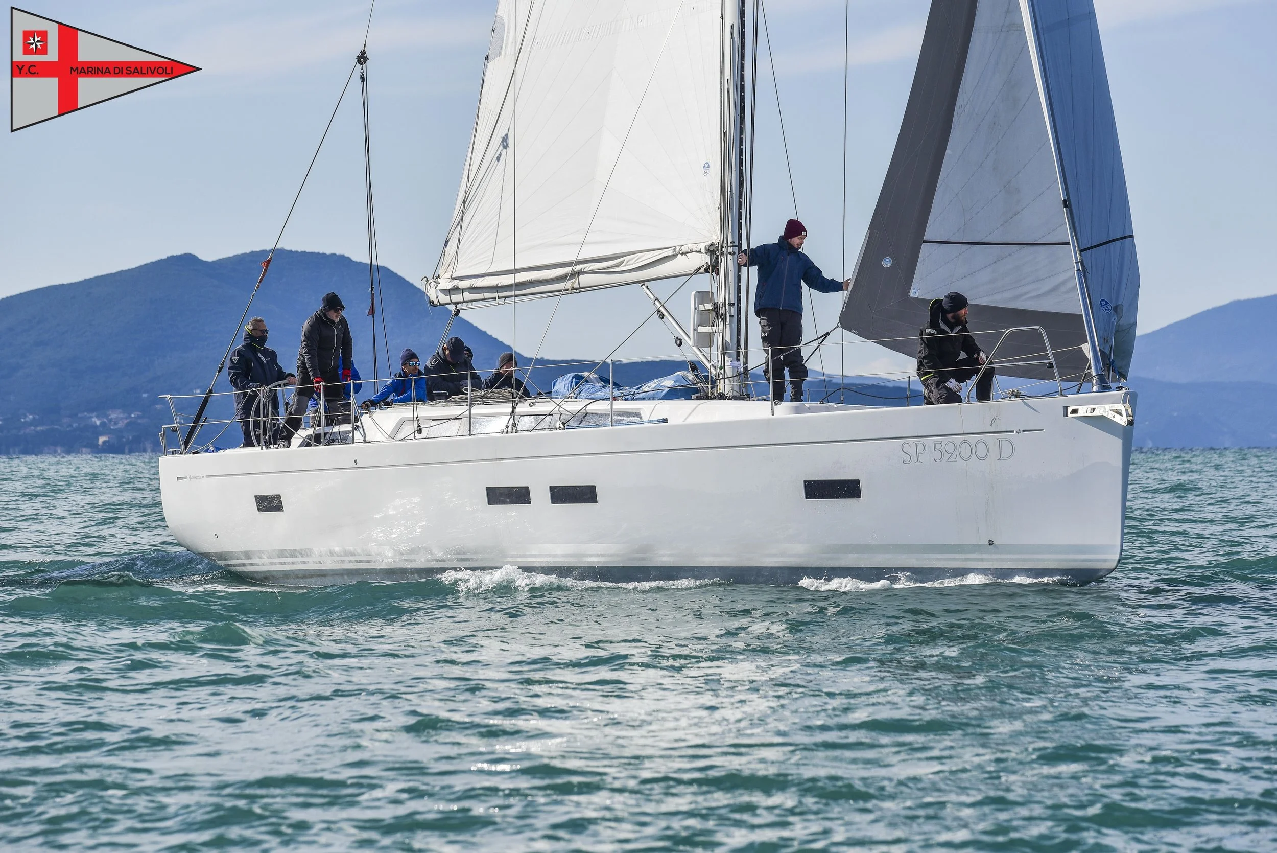 Yacht con equipaggio che naviga su acque azzurre con montagne sullo sfondo.