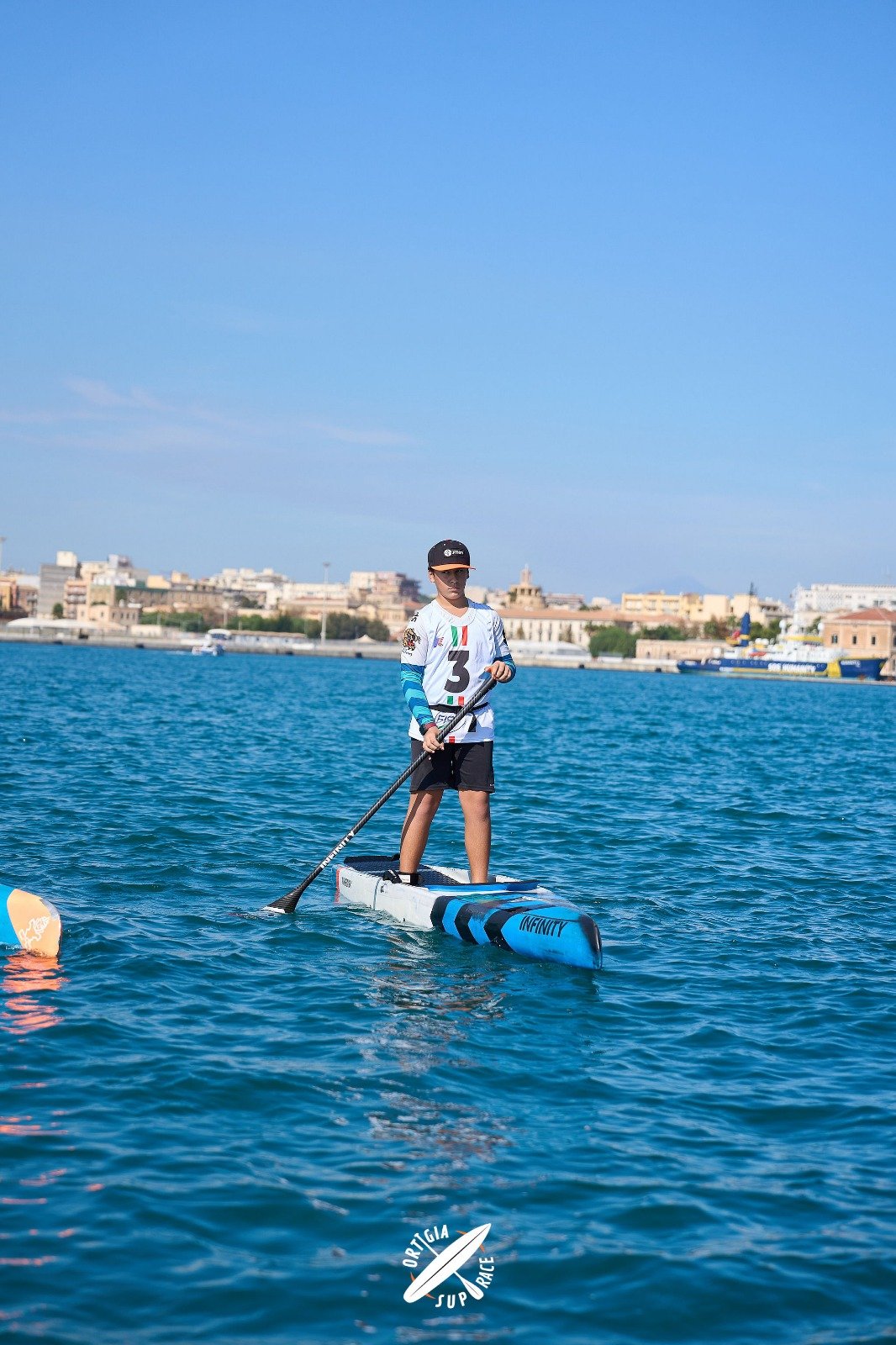 Un ragazzo giovane che si trova su una tavola da stand-up paddle sul mare, con una città sullo sfondo, indossa una maglietta bianca, pantaloncini neri, un cappellino nero e occhiali da sole.