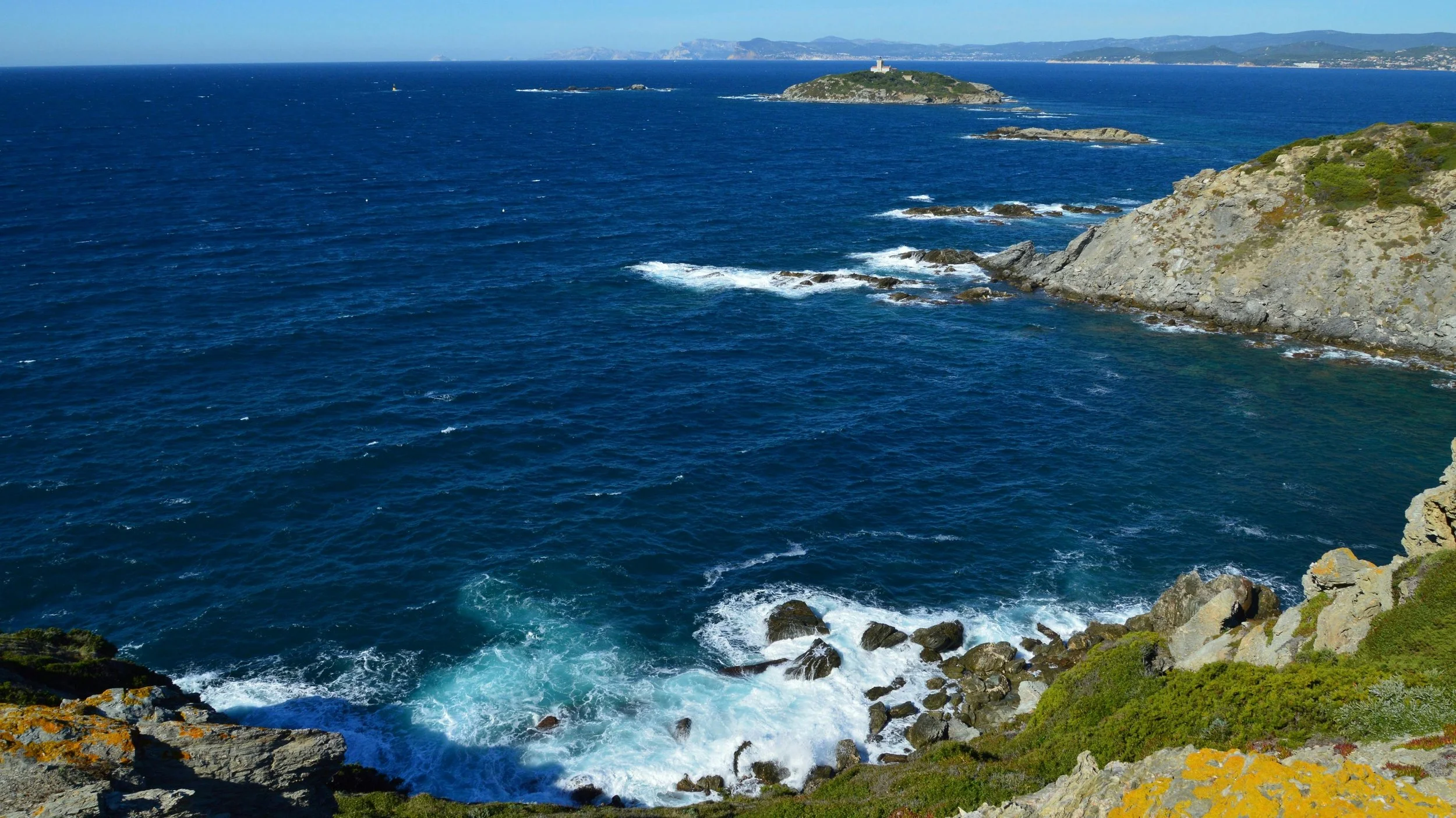 Costa rocciosa con onde che si infrangono e isole nel mare, con paesaggio montano in lontananza.