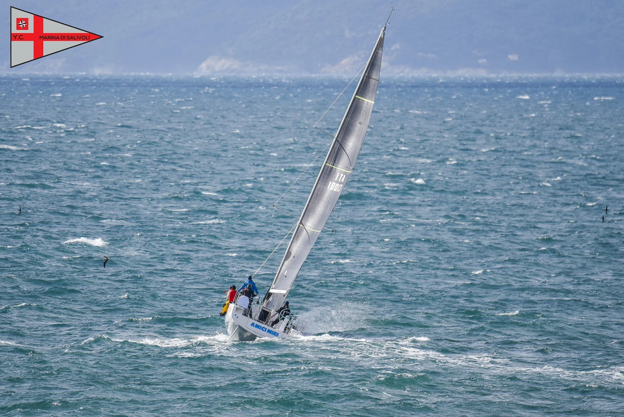 Una barca a vela inclinata a causa del vento in mare aperto con uno sfondo di cielo e costa lontana