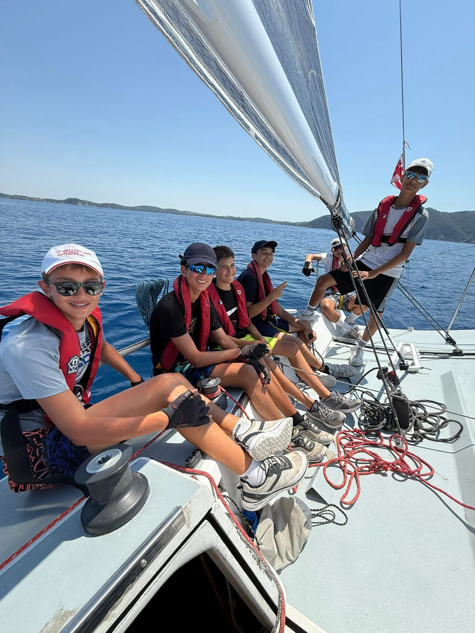 Gruppo di ragazzi seduti e in piedi sulla barca a vela, con alcuni che sorridono e fanno segno di vittoria, in acqua con paesaggio di isole sullo sfondo, giornata soleggiata.
