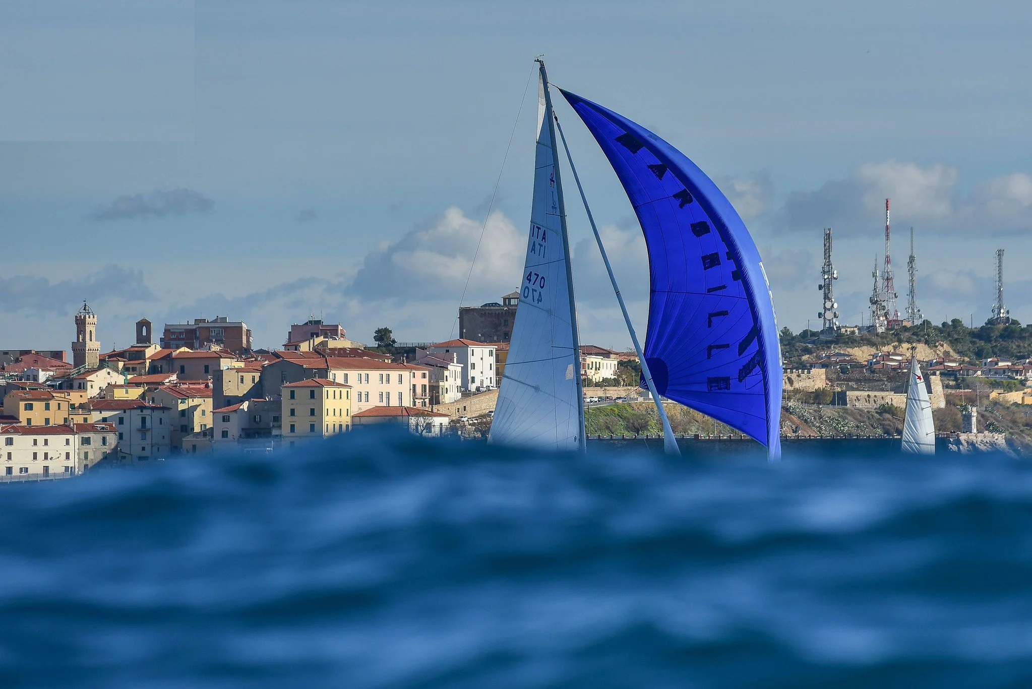 Una barca a vela con una grande vela blu mostra delle lettere nere, naviga vicino a una città con edifici multicolori e ricevitori radio su una collina, sotto un cielo nuvoloso.
