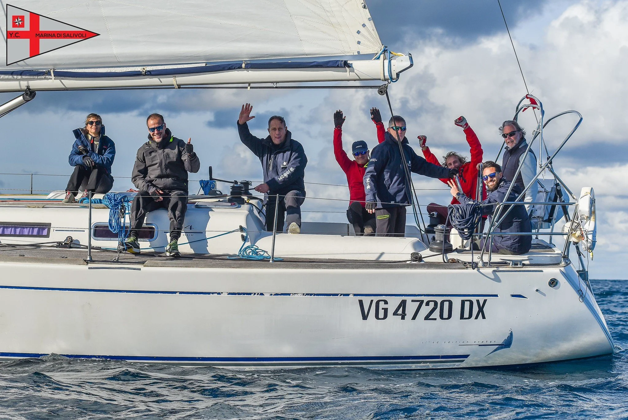 Gruppo di persone su una barca a vela, sorridenti e che salutano, con un cielo nuvoloso sul mare.