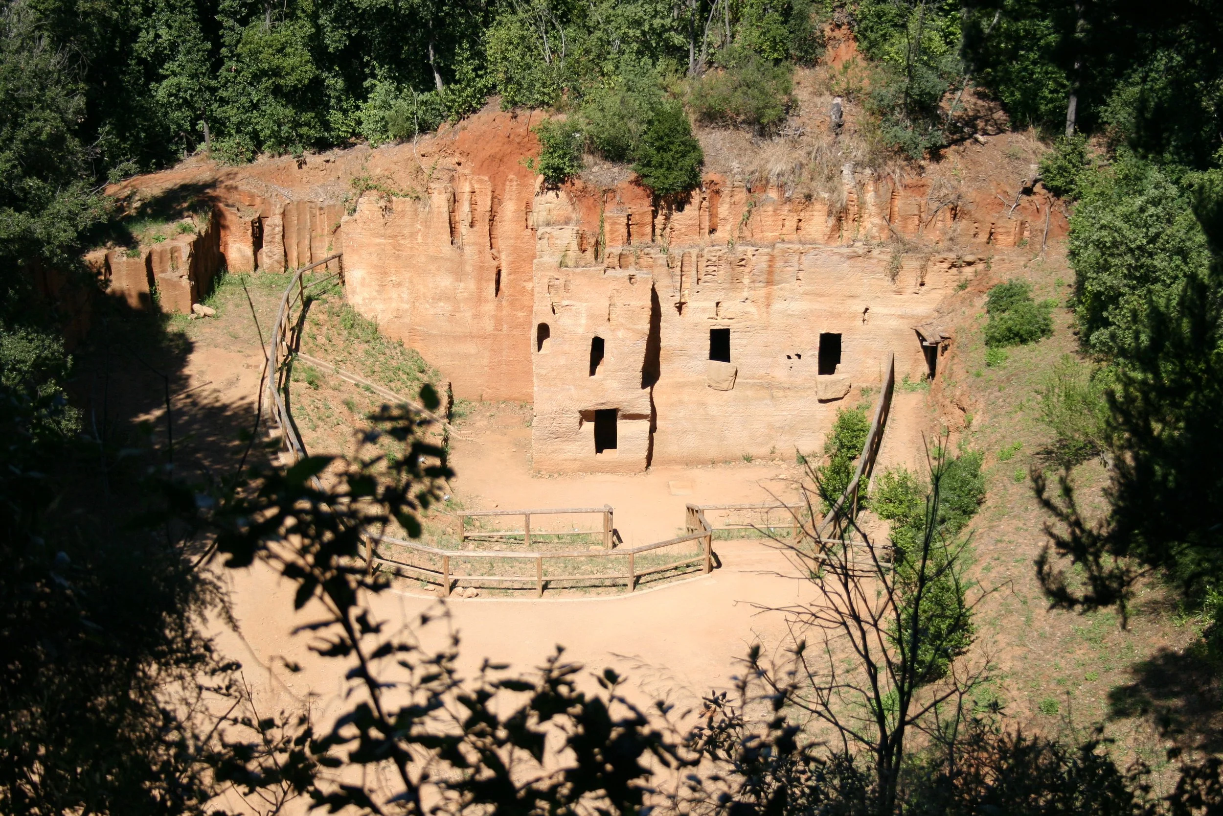 Scavi archeologici di una antica abitazione in pietra rossa, circondata da vegetazione e alberi.