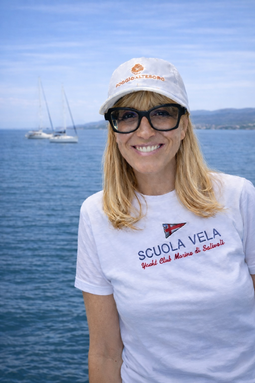 Una donna con cappello bianco, occhiali neri, sorriso e maglietta bianca con scritte e logo, sullo sfondo un mare con barche e un cielo sereno.