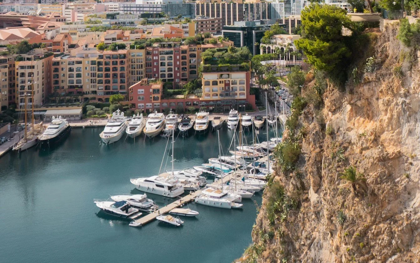 Porto con yacht e barche, davanti a edifici residenziali e un pendio roccioso con vegetazione.