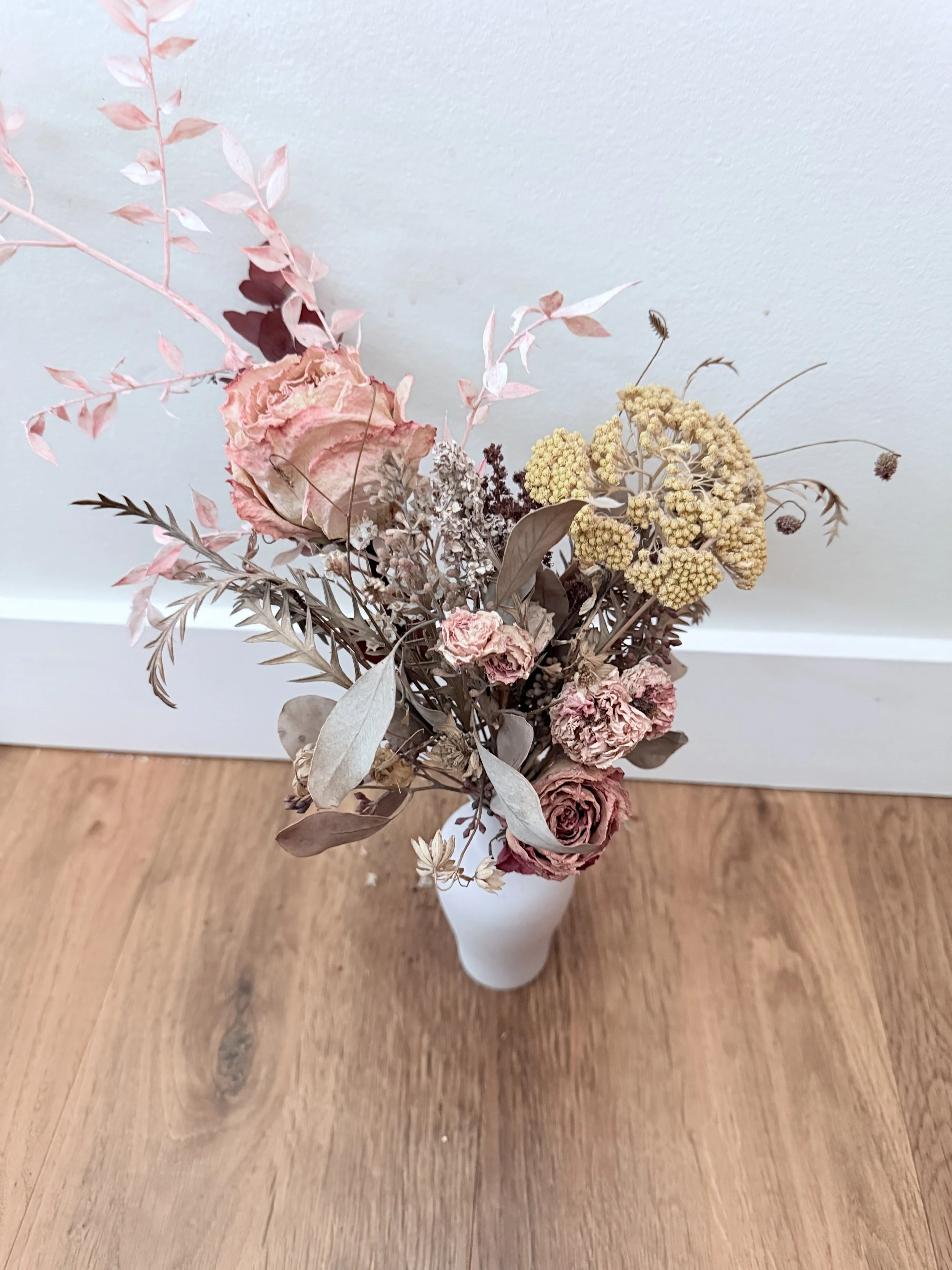 A dried flower bouquet in a white vase on a wooden table, featuring pink roses, beige and brown foliage, and yellow and purple accent flowers, they form a heart shape.